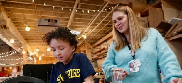An adult with a lanyard badge sits beside a young child working at a table indoors.