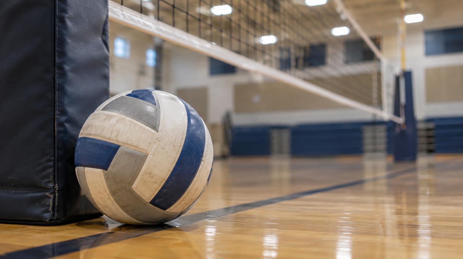 A volleyball resting on a gym floor next to a volleyball net post.
