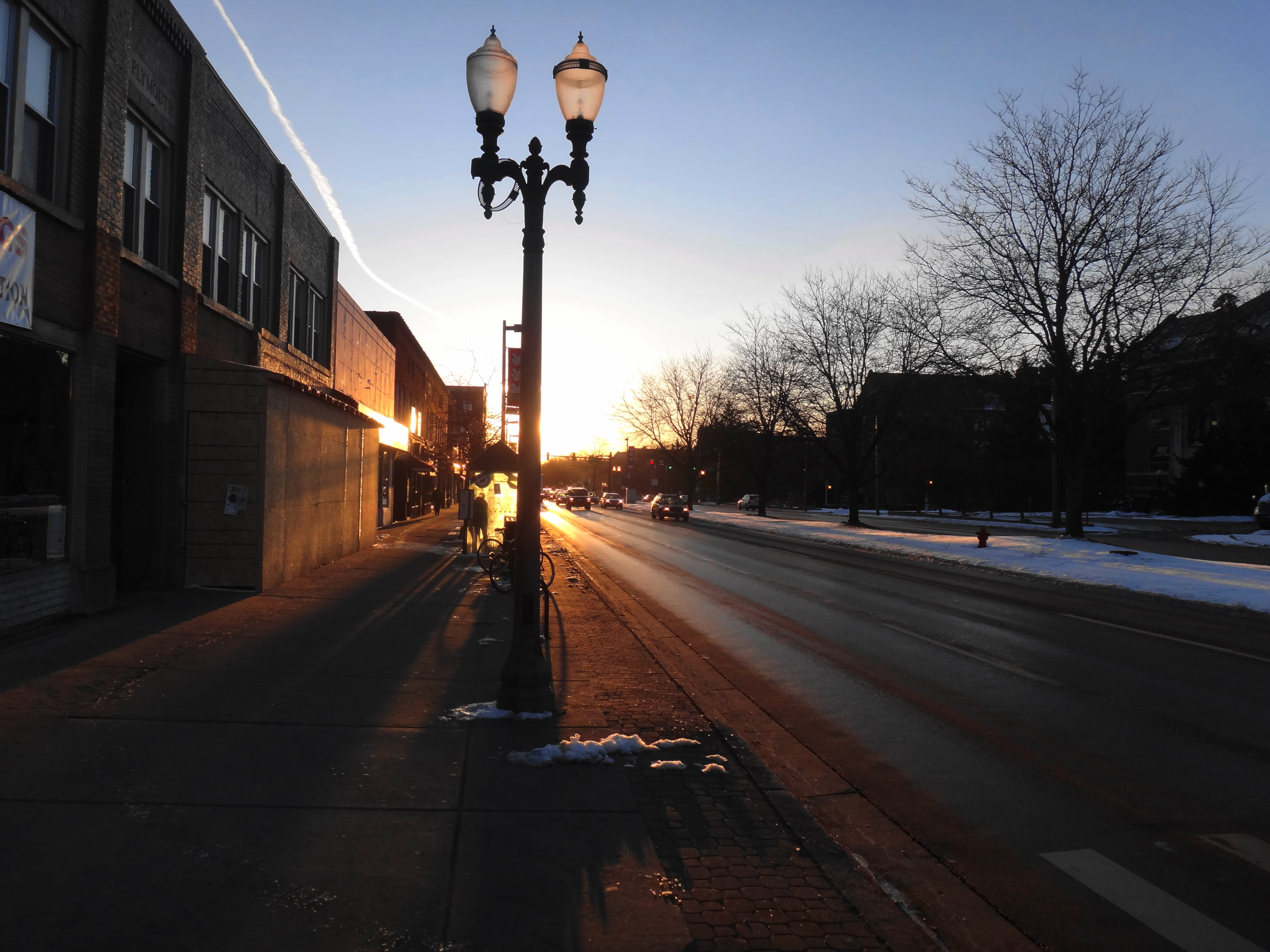 Street lamp on a snowy sidewalk along a wide avenue at sunrise, brick buildings visible in the distance.