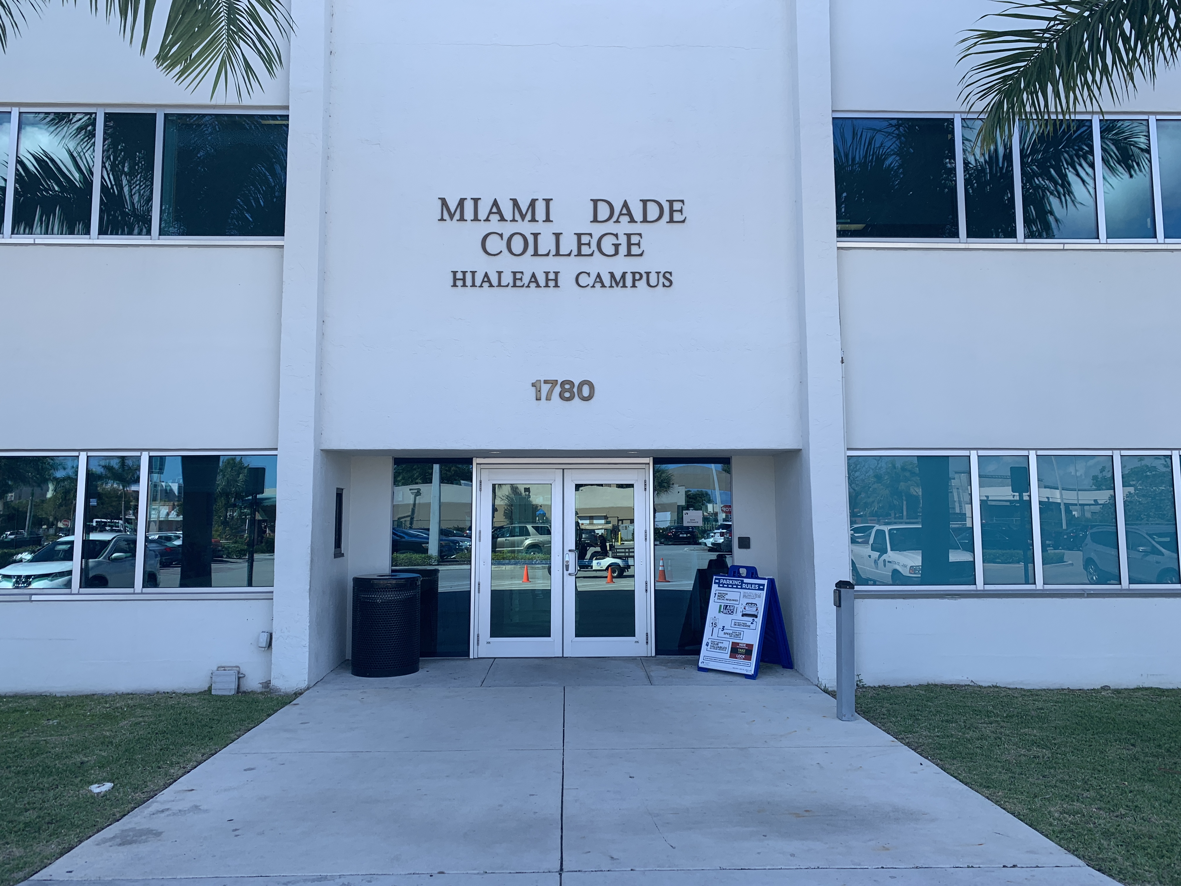 Glass entrance doors of a white multi-story Miami Dade College building flanked by palm trees.