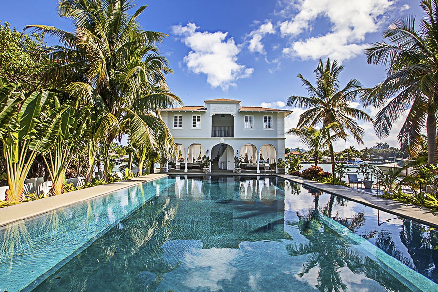 White Mediterranean-style mansion with a large rectangular pool and surrounding palm trees.