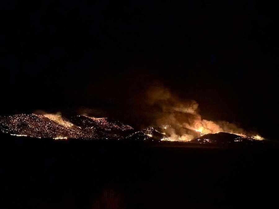 A wildfire burns across multiple hillsides at night with flames and smoke illuminating the dark sky.