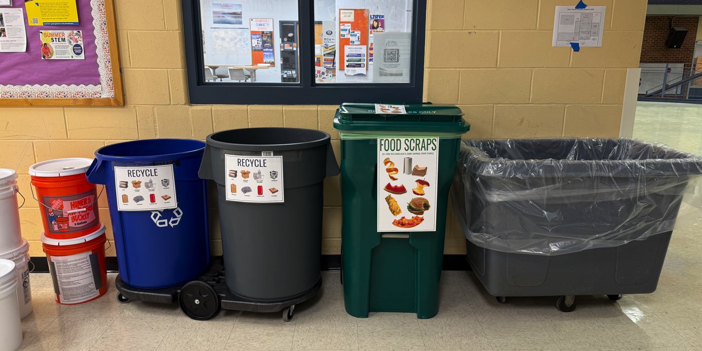 Four color-coded waste-sorting bins lined up against a yellow-brick school hallway wall.