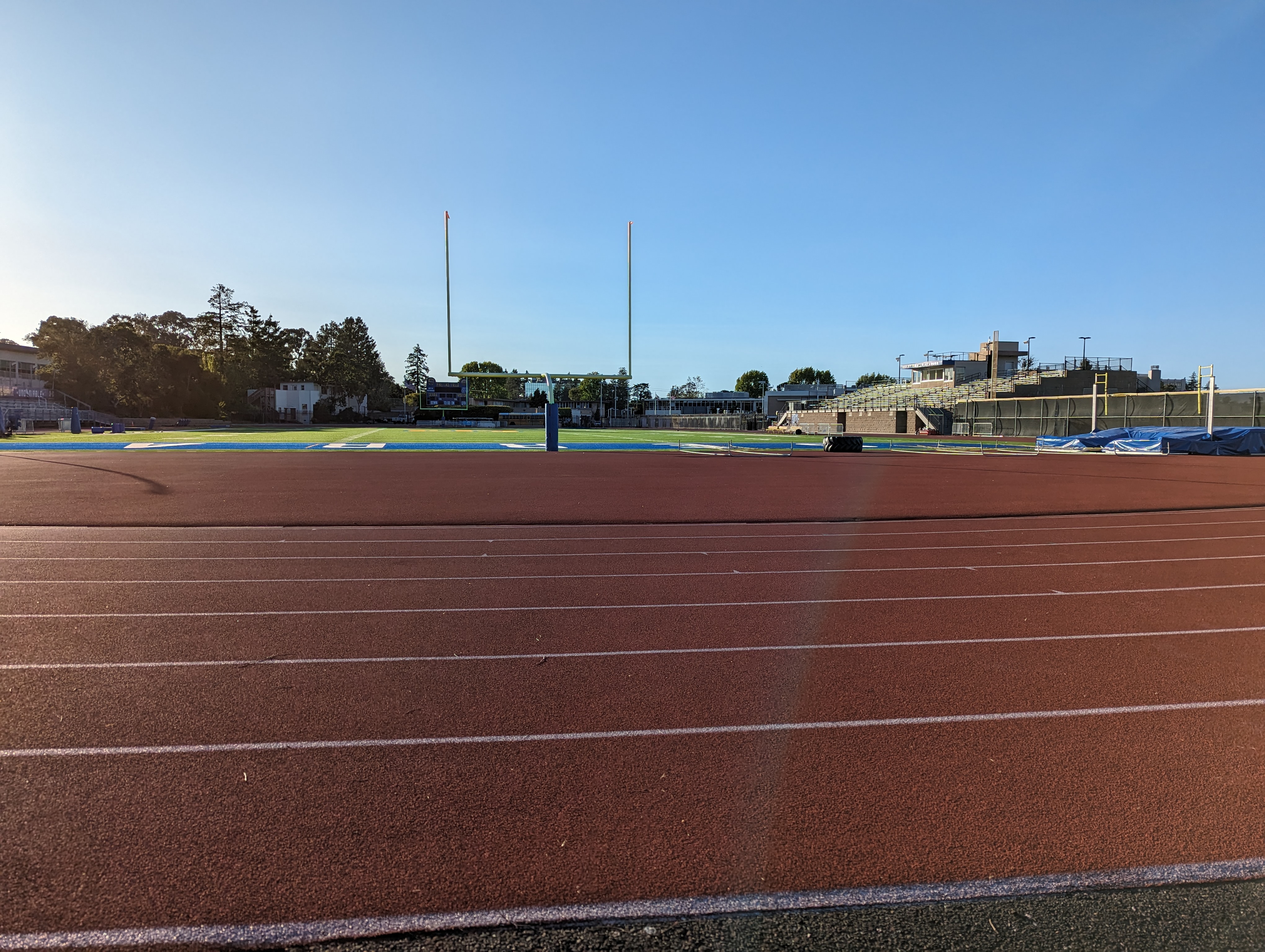 Red running track and football field with goalposts, bleachers, and scoreboard under blue sky.