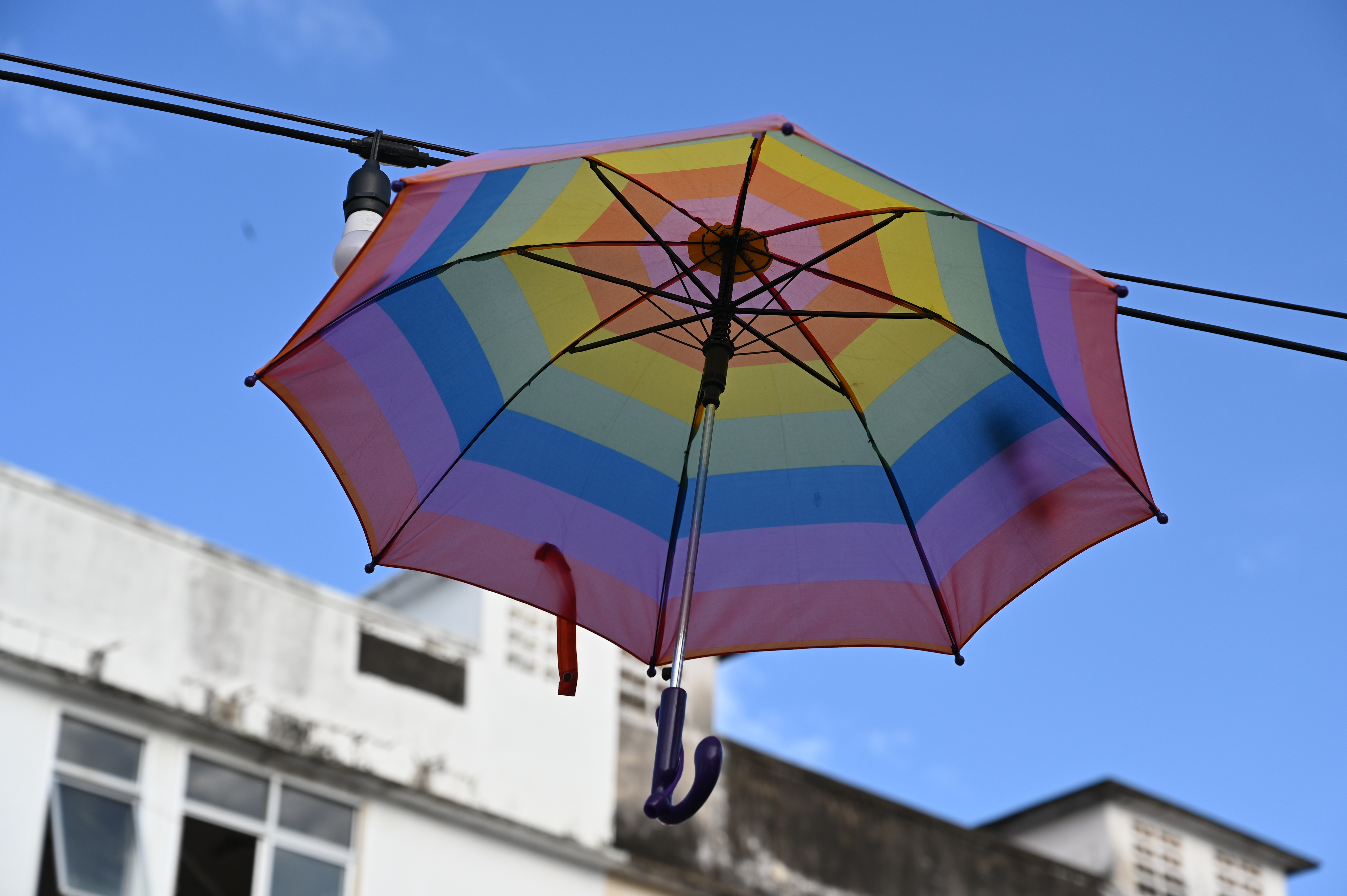 A vibrant rainbow-colored umbrella suspended from a wire against a clear blue sky.