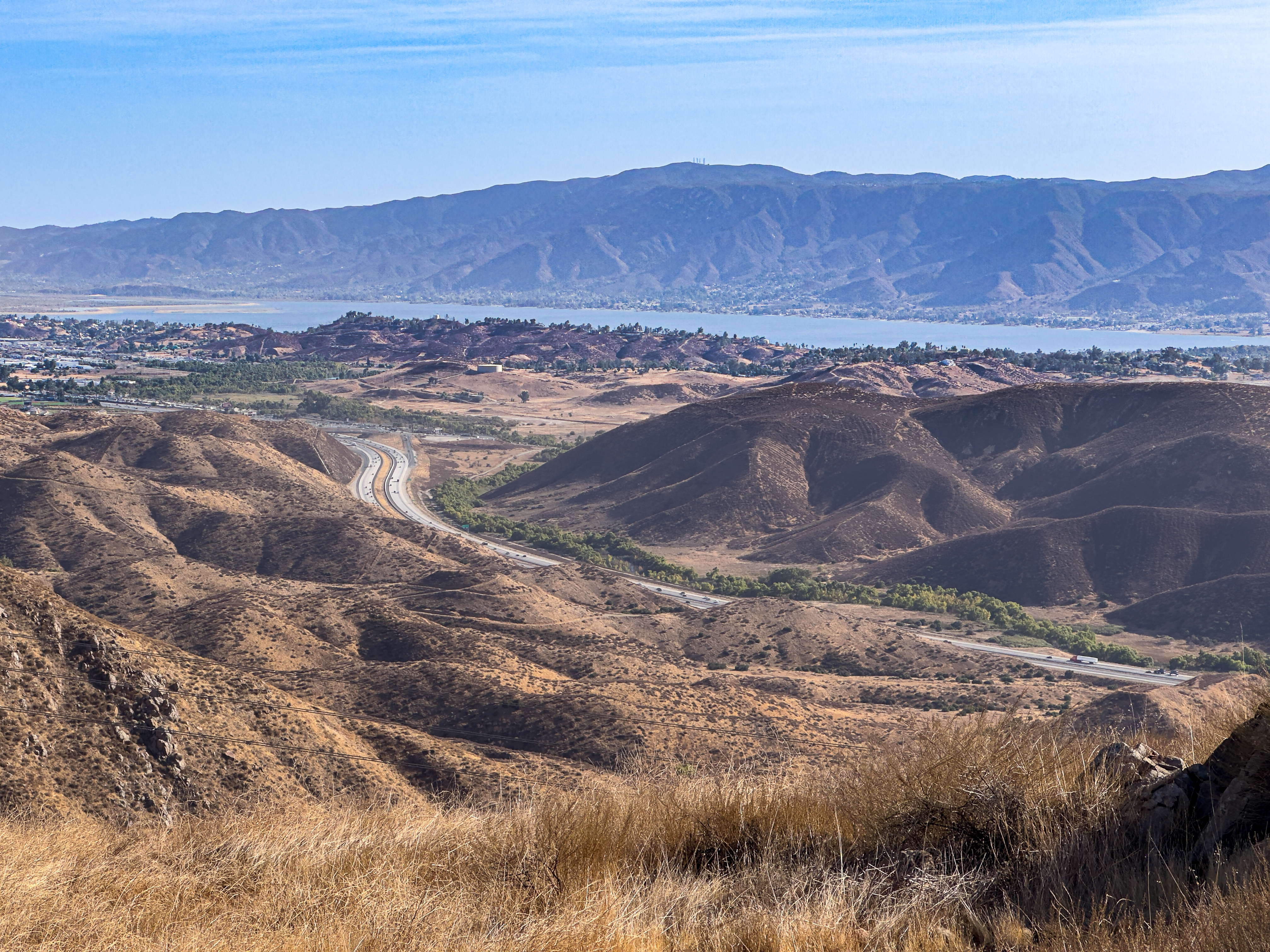Multi-lane freeway cutting through dry chaparral hills with a lake and valley in the background.