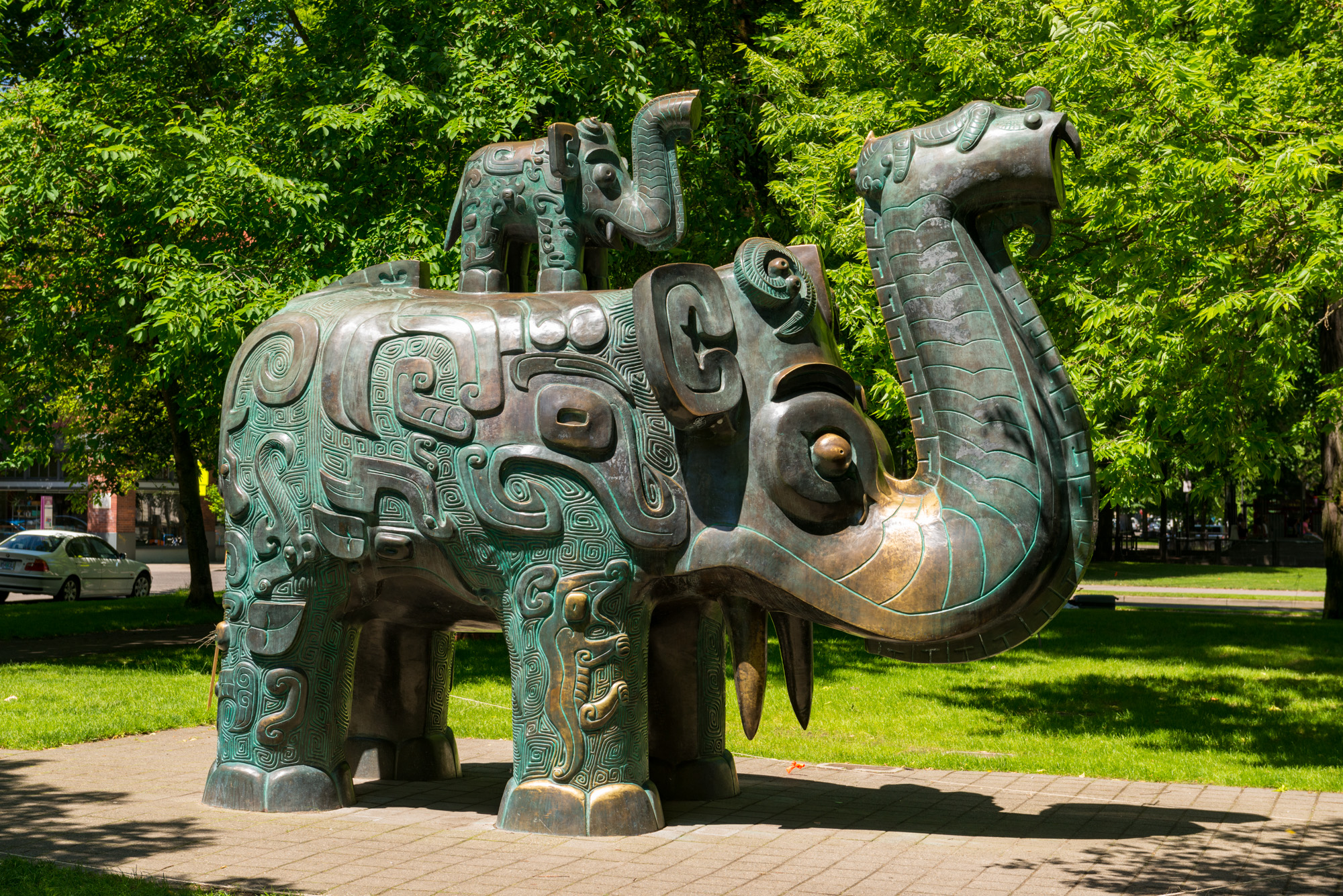 Large ornate bronze elephant sculpture on a paved path surrounded by park trees.