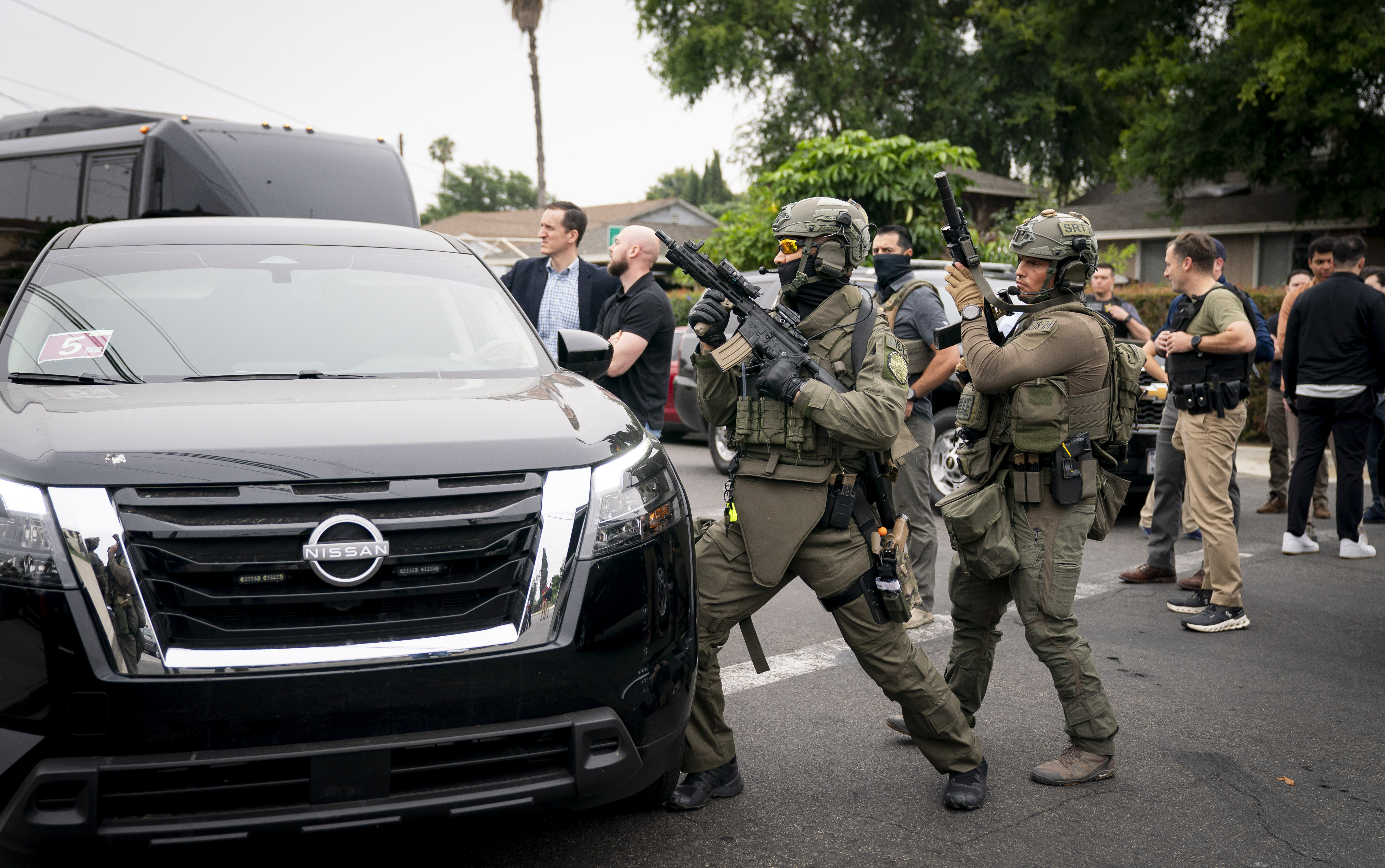 Two tactical officers in green gear approach a black SUV on a residential street.