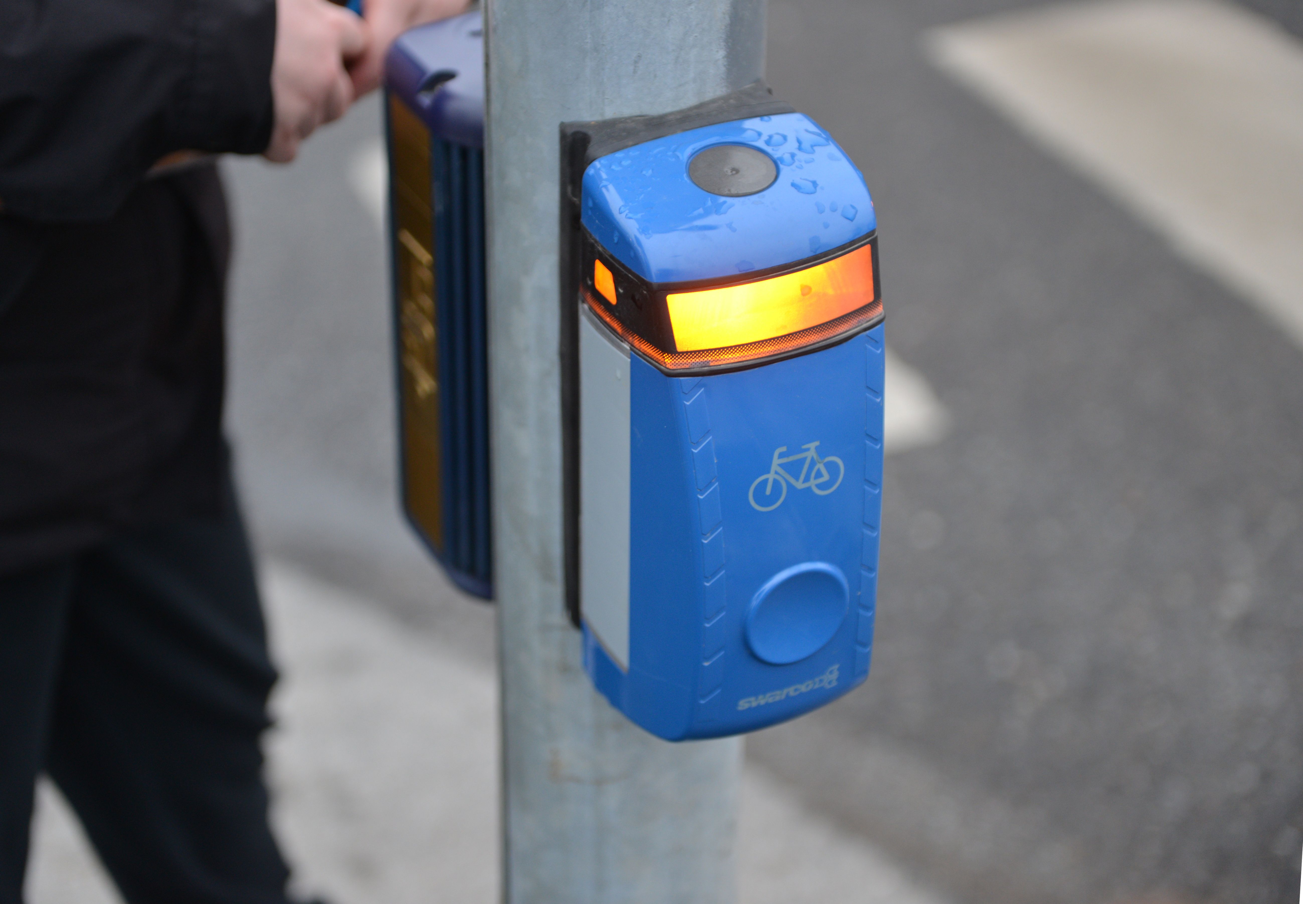 Blue push-button pedestrian and bicycle crossing signal on a metal pole with amber light lit.