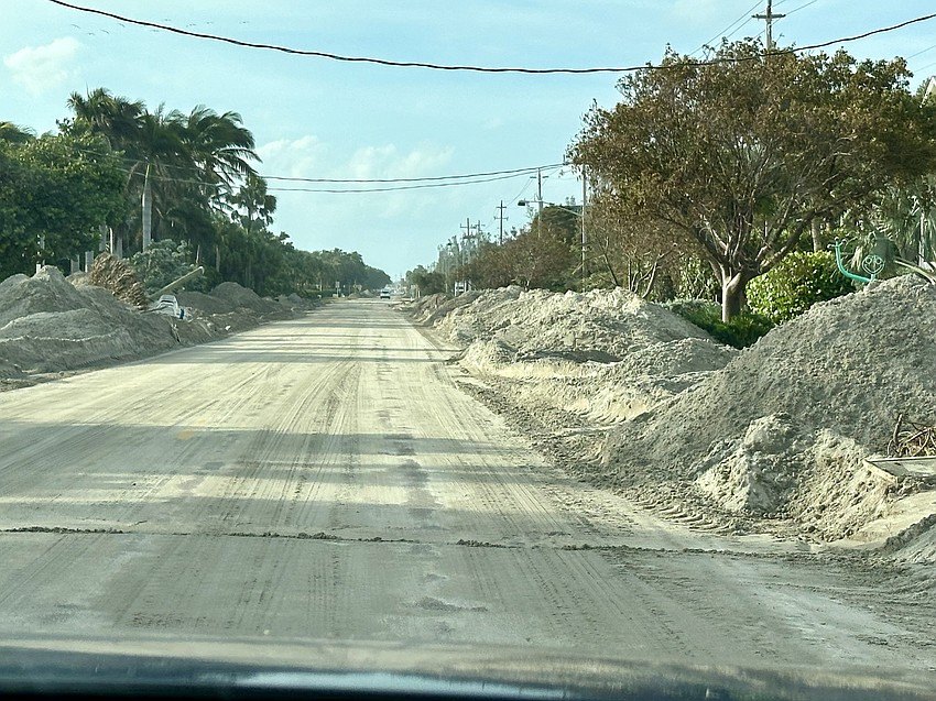 A road flanked by large sand and debris mounds, with palm trees and utility lines visible.
