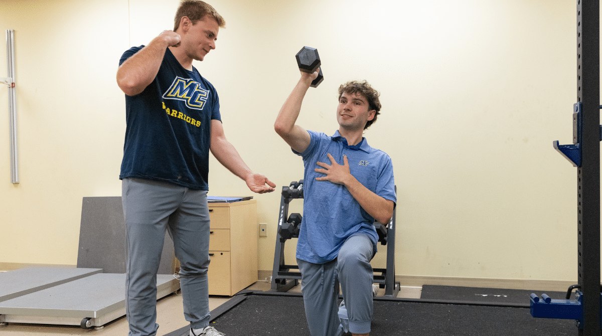 One person in a Merrimack shirt watches another perform a kneeling dumbbell press in a gym.