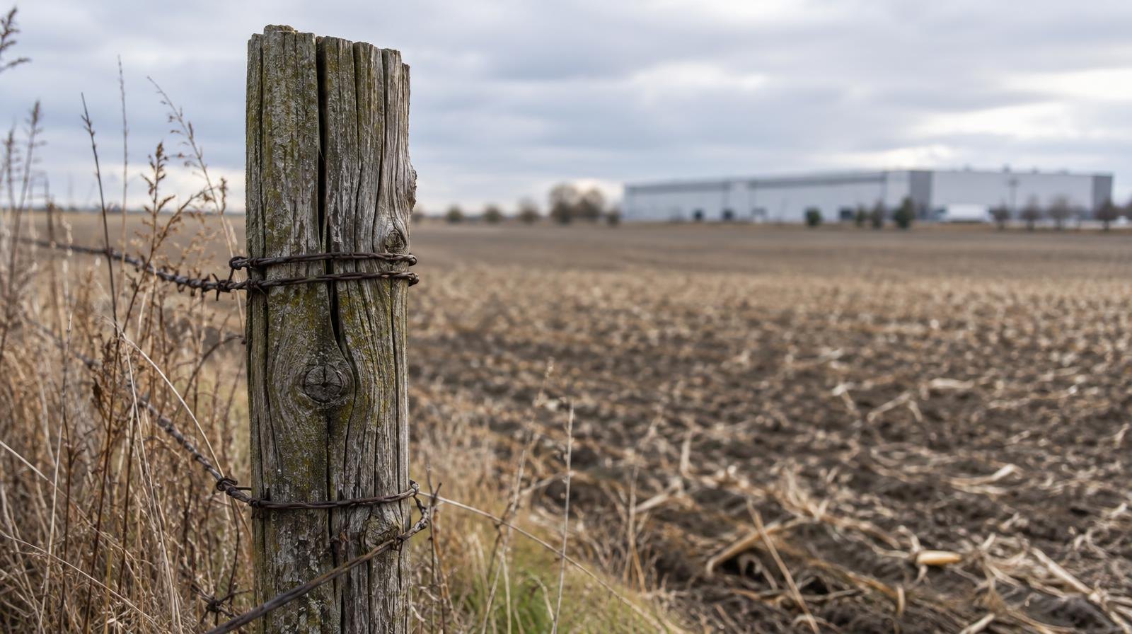 Fallow farm field with a wooden fence post and distant warehouse on the horizon.