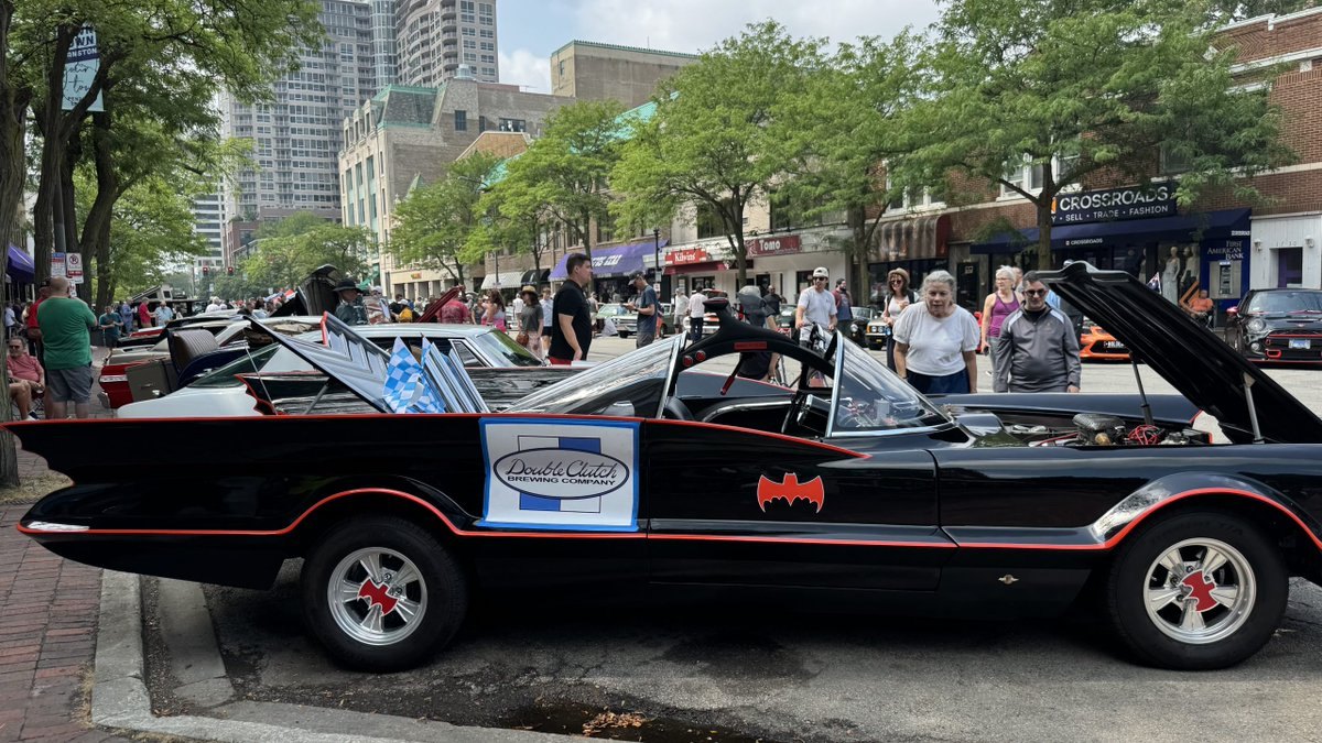 Black replica Batmobile with red trim and fins parked on a city street at an outdoor car show.