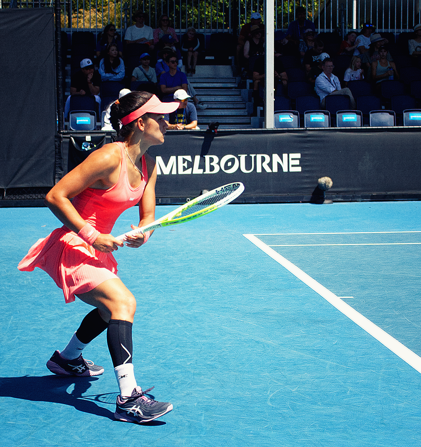 Female tennis player in pink dress and visor ready to return a shot on a blue hard court.