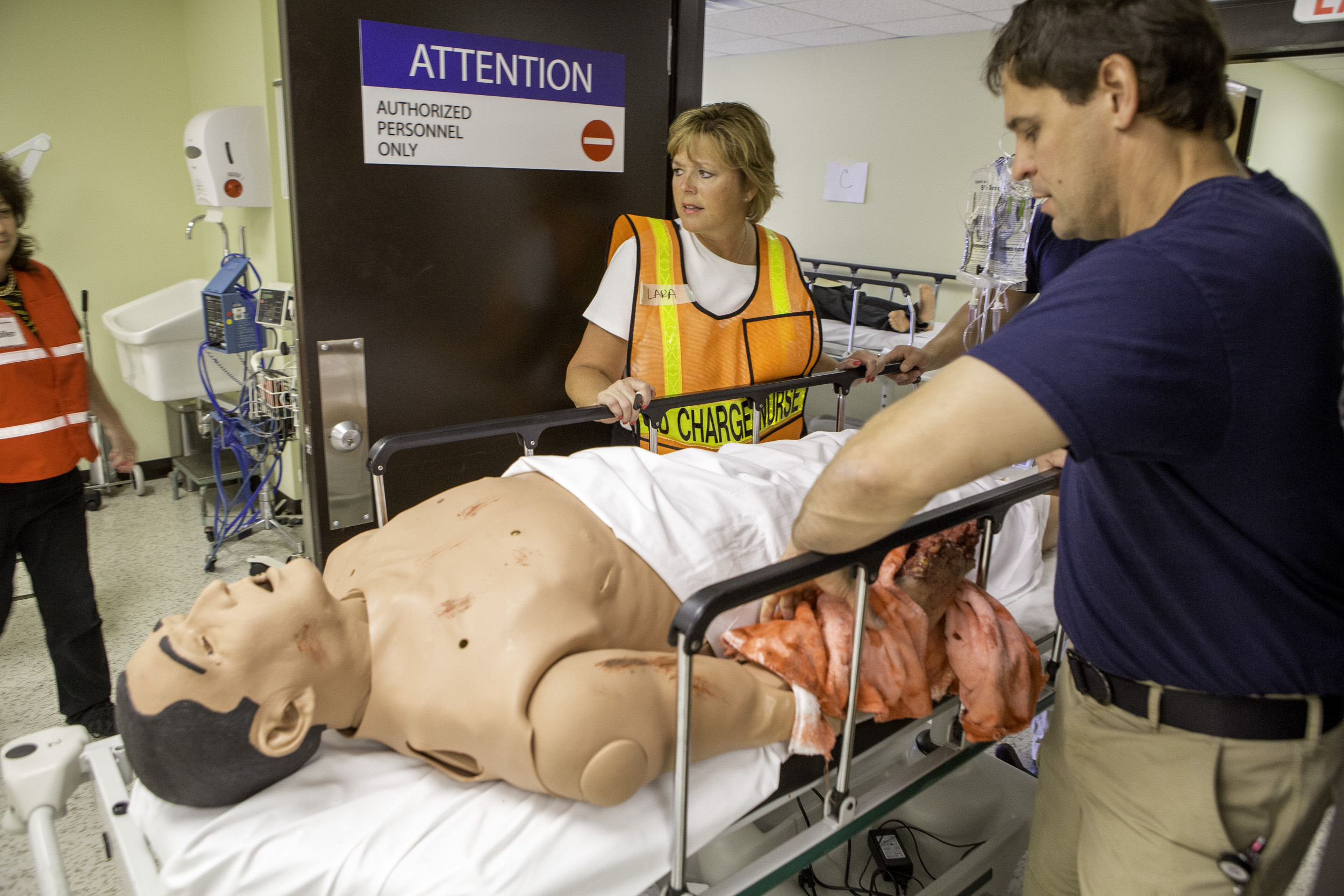 Emergency responders wheel a mannequin on a gurney through a medical facility hallway.