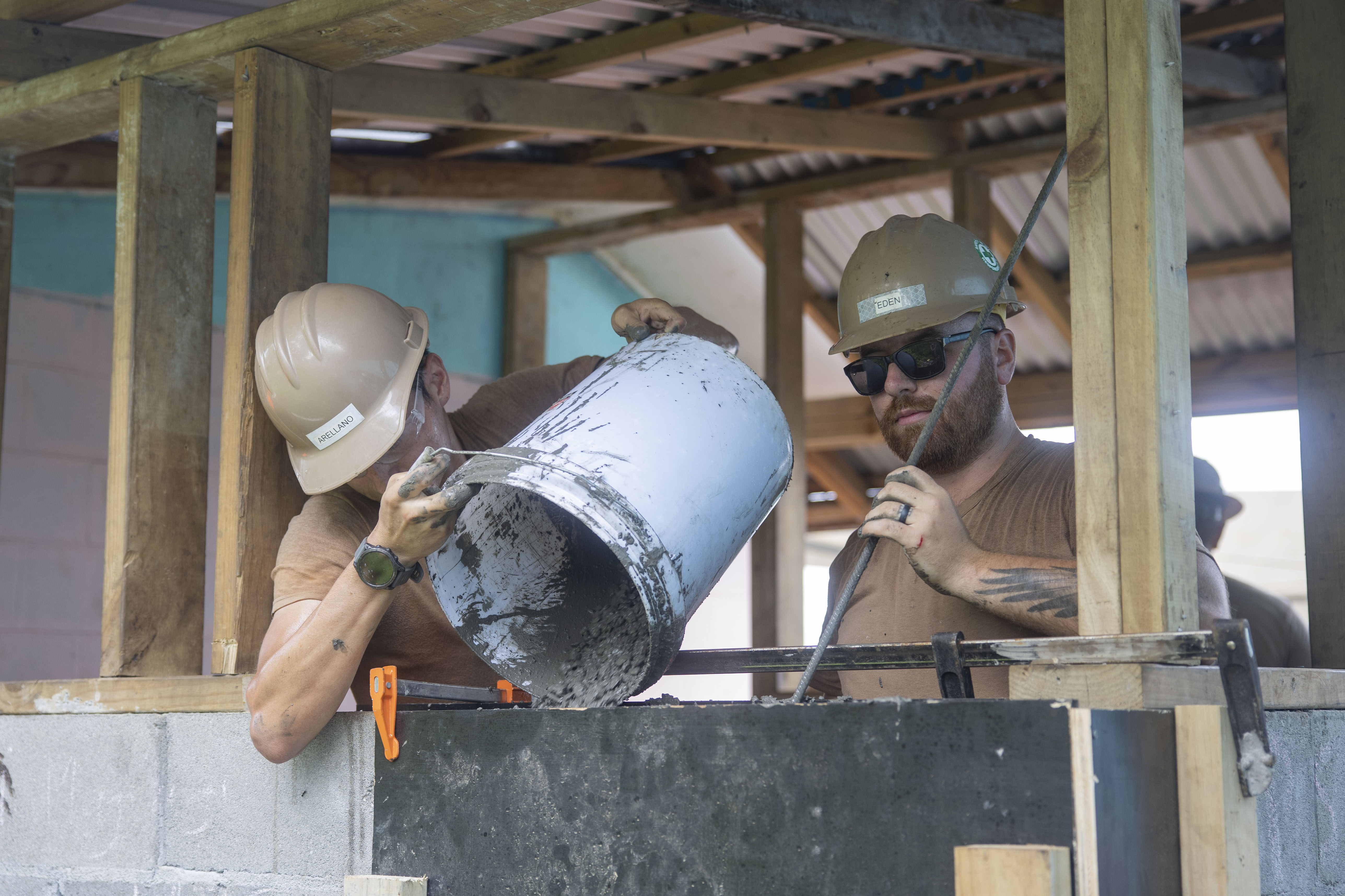 Two workers in hard hats at a construction site, one pouring material from a bucket.