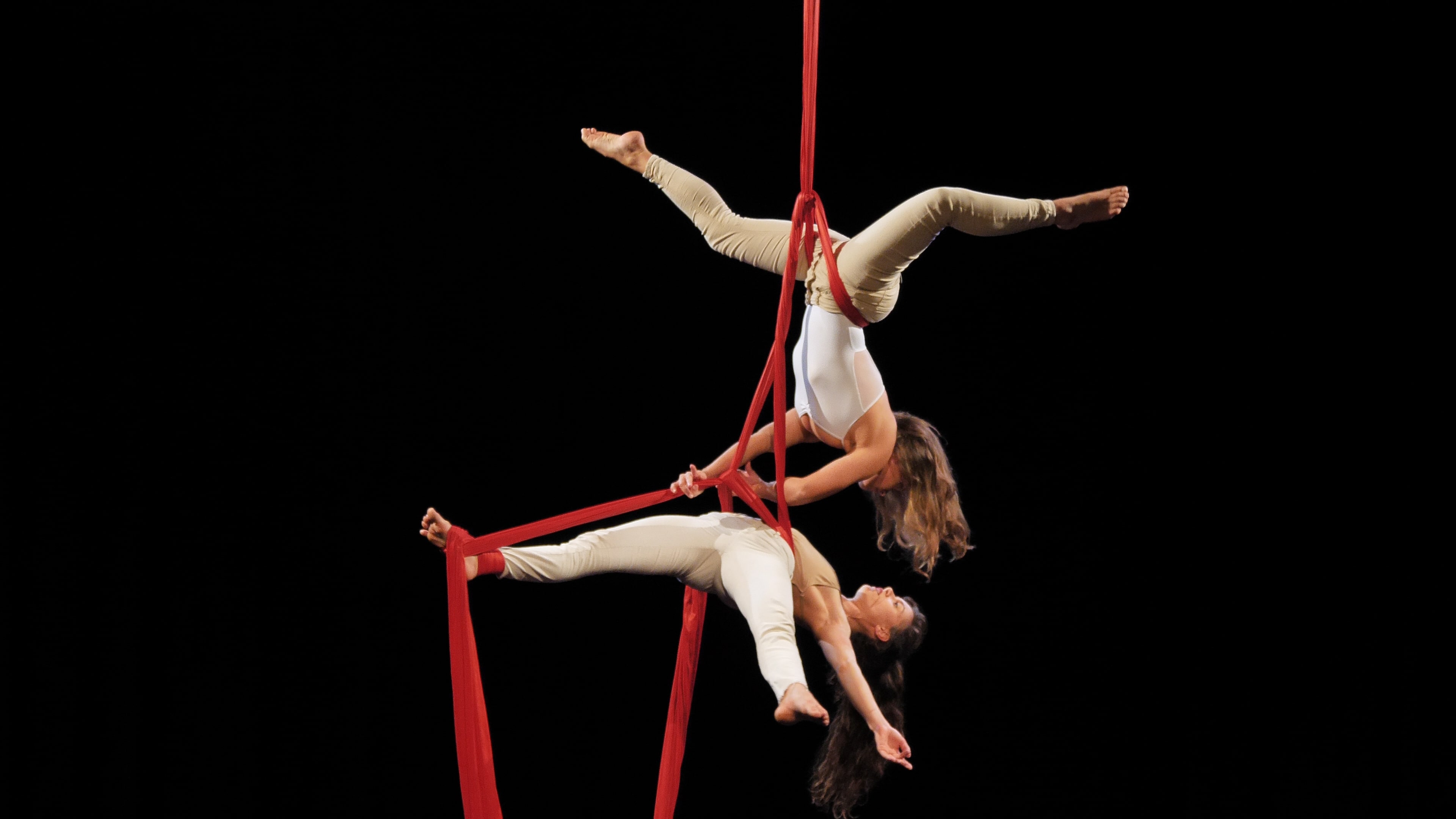 Two performers in white costumes suspended on red aerial silks.