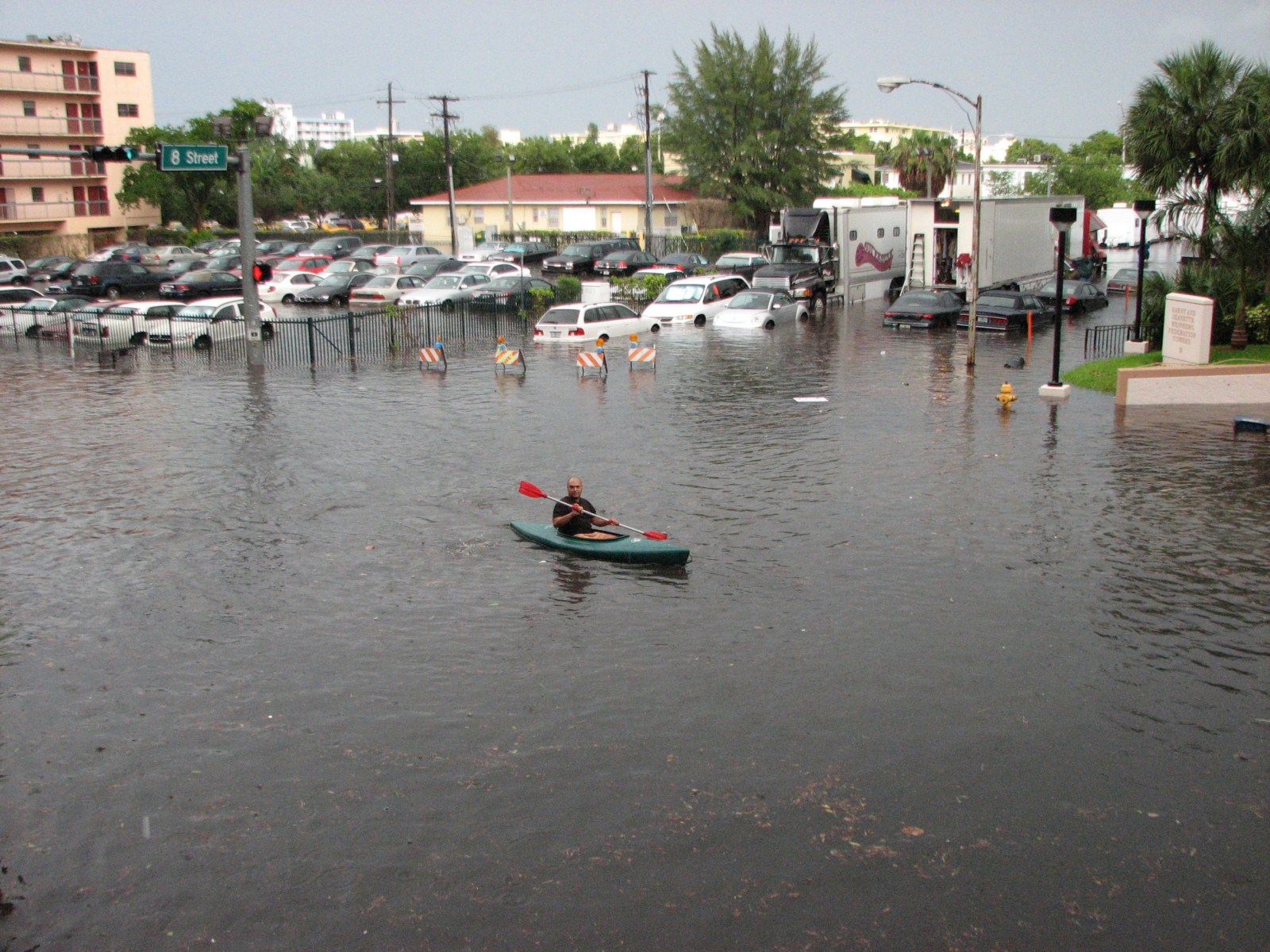 A kayaker paddles through a flooded Miami Beach street intersection with submerged cars nearby.