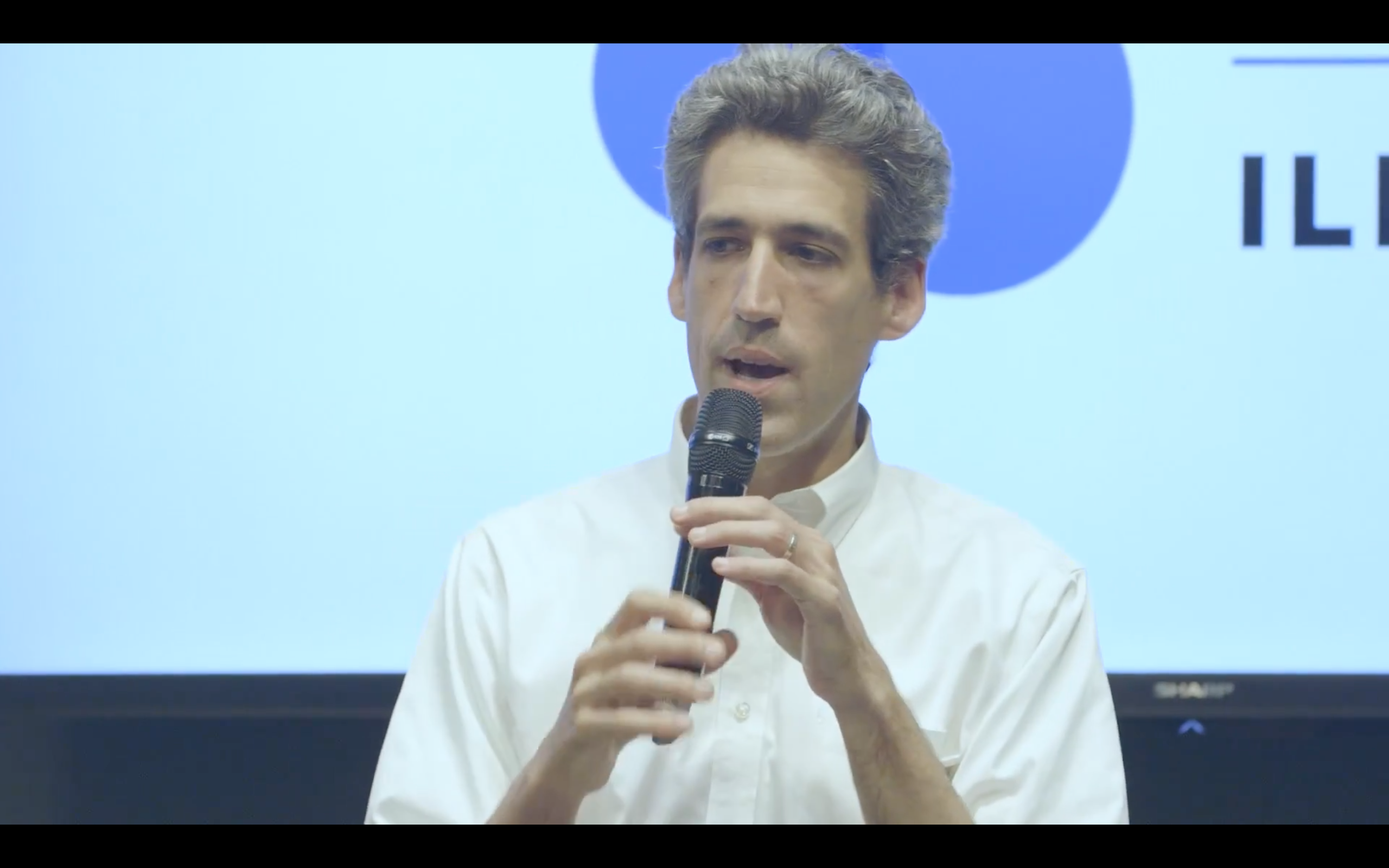 A man with gray hair speaking into a handheld microphone in a white button-down shirt.