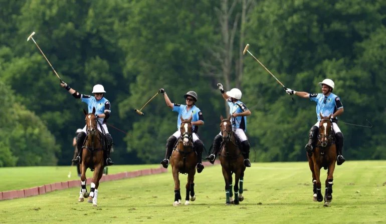 Four polo players in light blue jerseys on horseback raising mallets in celebration on a green field.