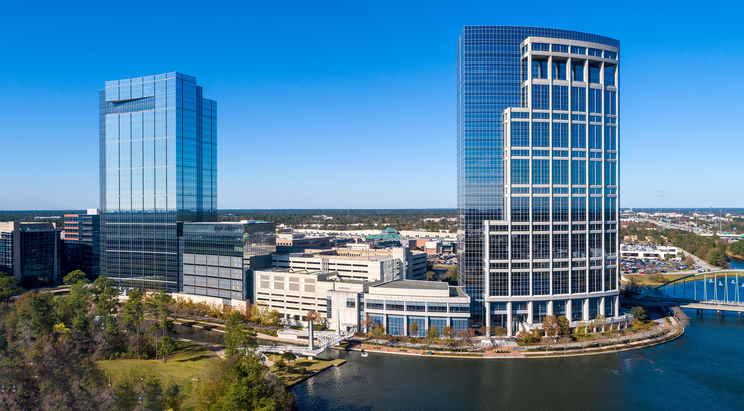 Aerial view of two tall glass towers along a waterway with trees and commercial buildings below.