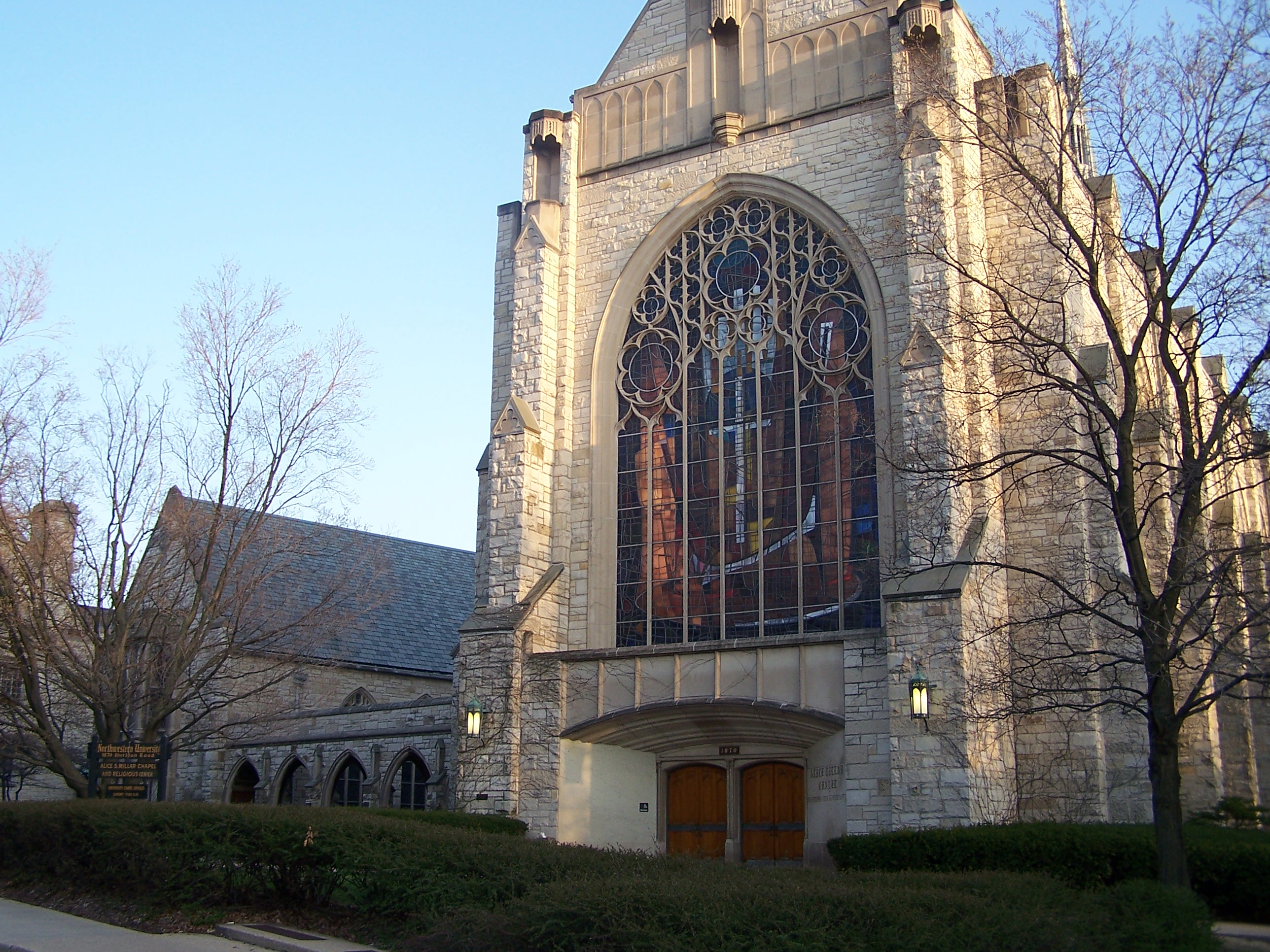 Gothic stone chapel with a large arched stained-glass window and bare trees.