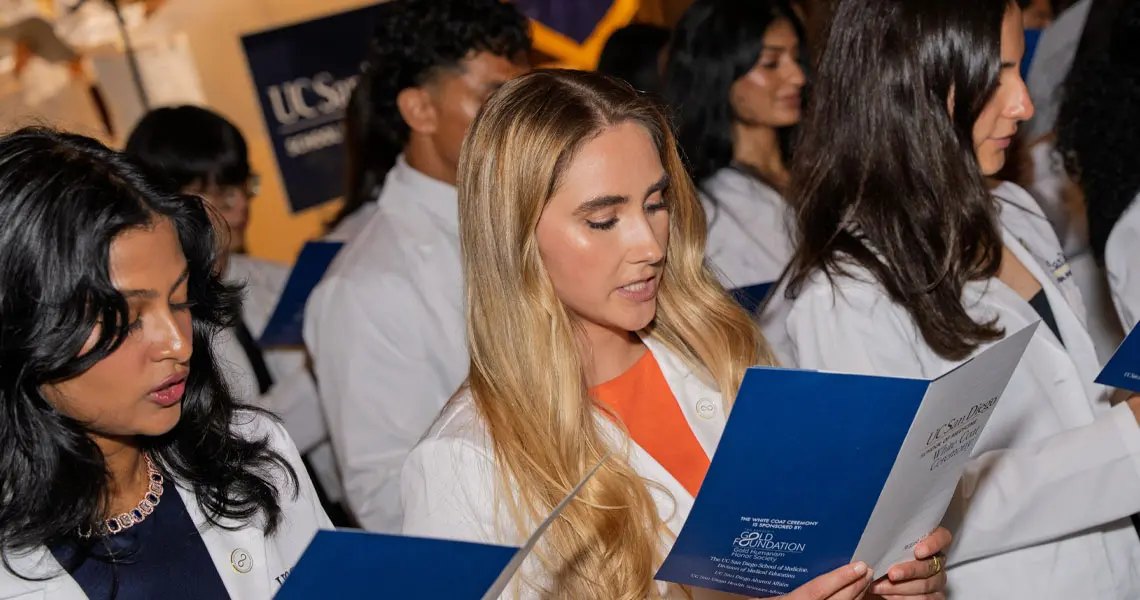 Three women in white coats read from blue event programs at an indoor ceremony, with more white-coated students behind them.