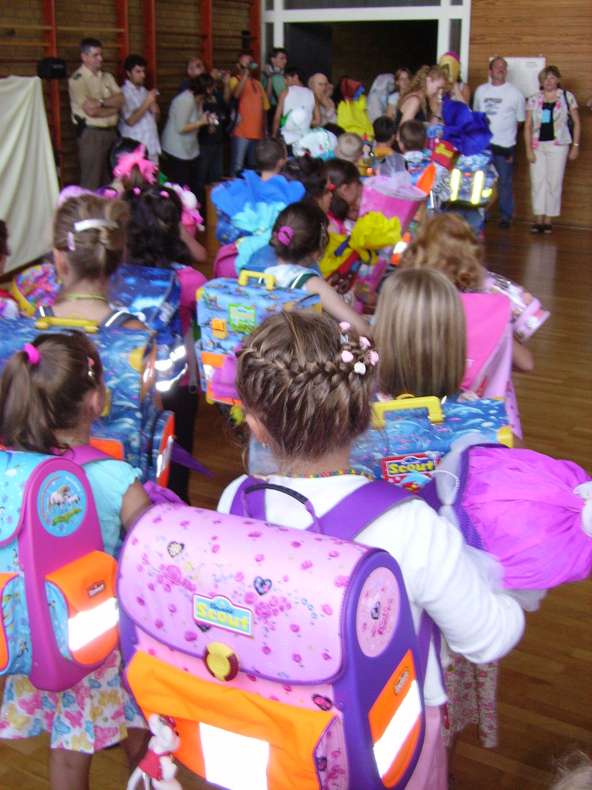 Young children wearing colorful backpacks gathered in a school hall, seen from behind.