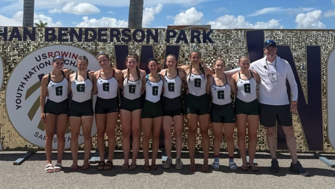 Youth women's rowing team in green and white uniforms posing in front of Nathan Benderson Park sign at nationals.