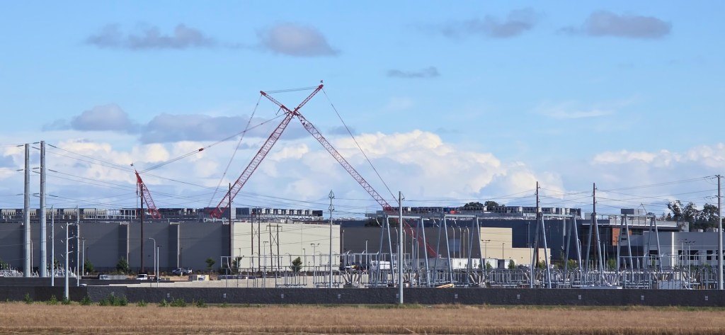 Large data center buildings with construction cranes behind electrical substation and power lines, viewed across an open field.