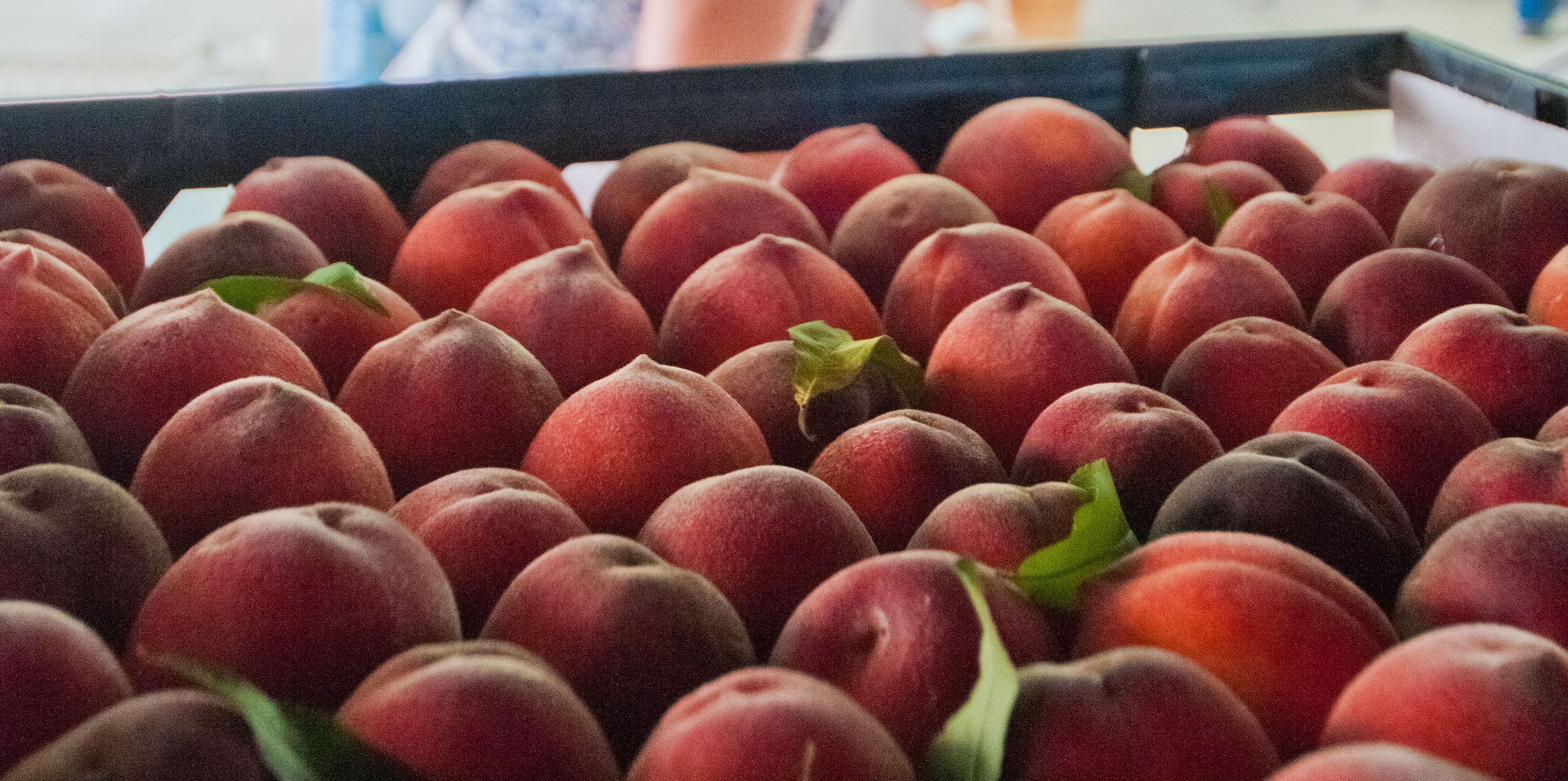 A crate of ripe red-orange peaches with green leaves at a farm market stand.