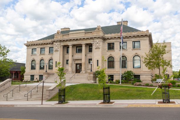 Neoclassical stone library building with columns, green roof, and American flag on a sunny day.