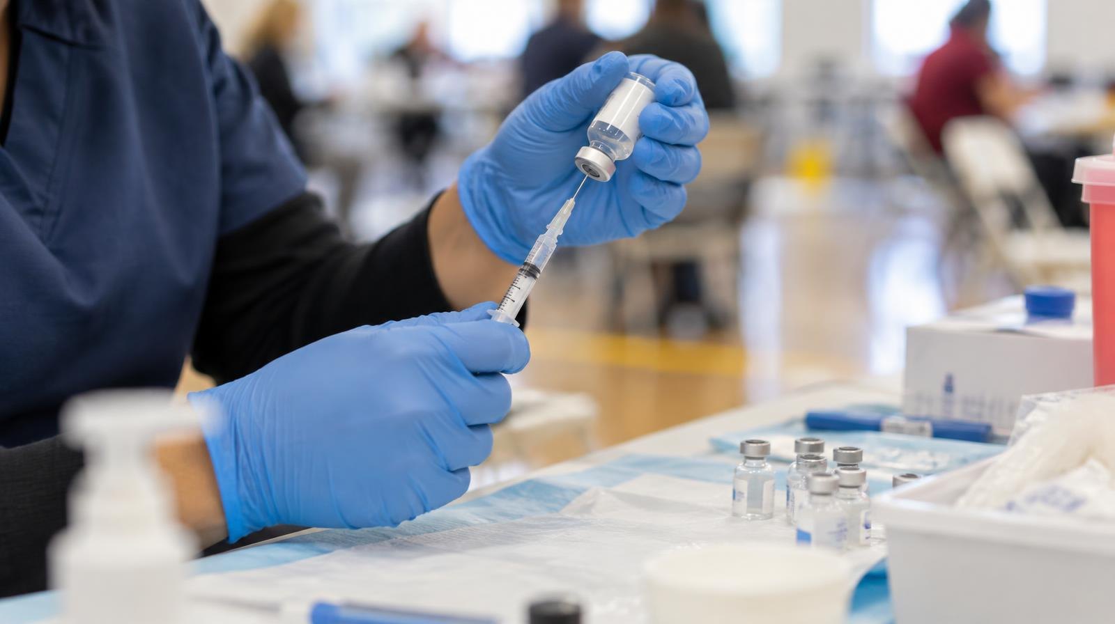 A gloved hand holds a syringe next to small vaccine vials on a tray.