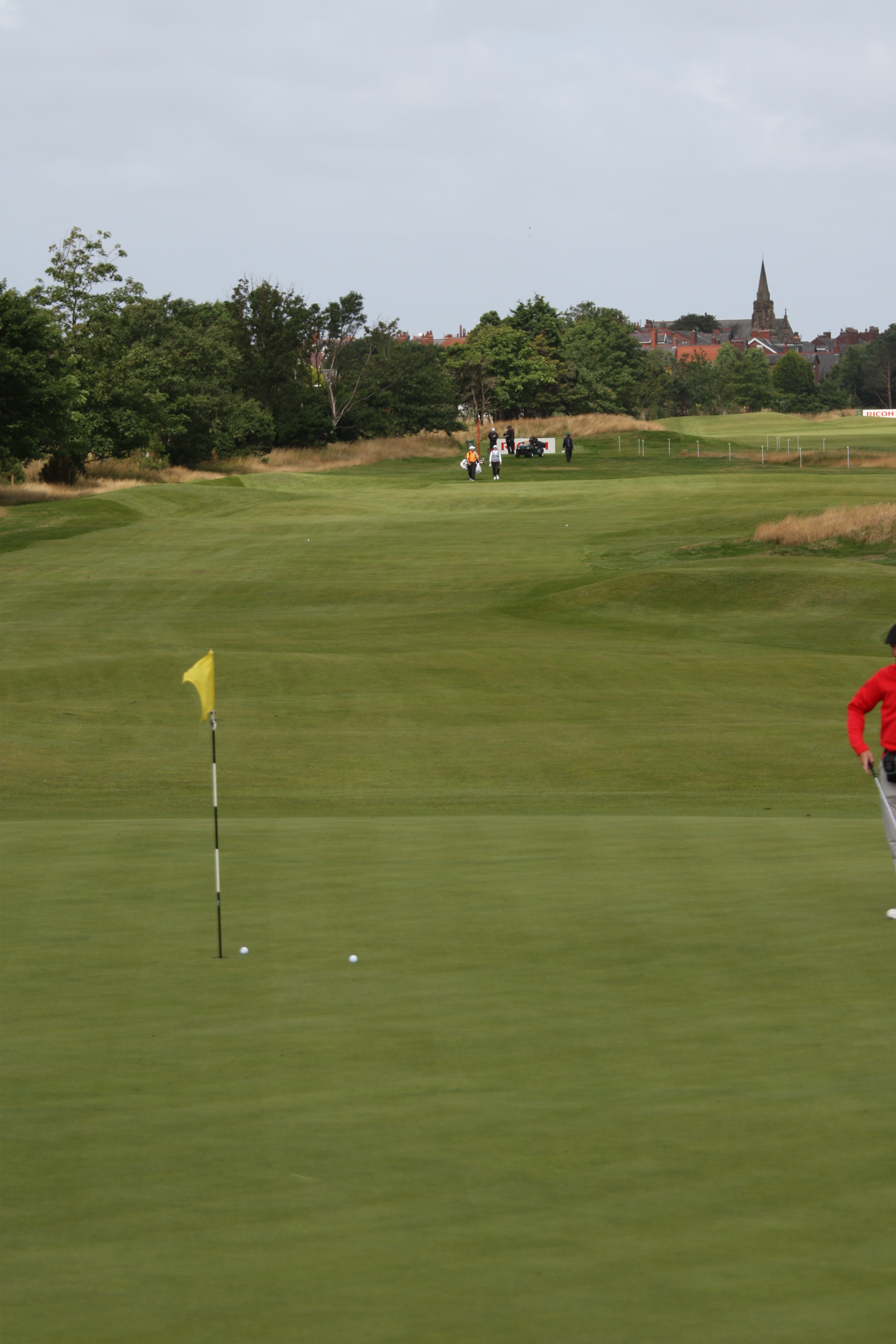 A golfer in red stands near a flagged hole on a putting green.
