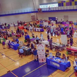 Overhead view of a busy indoor community fair with vendor tables and attendees in a gymnasium.