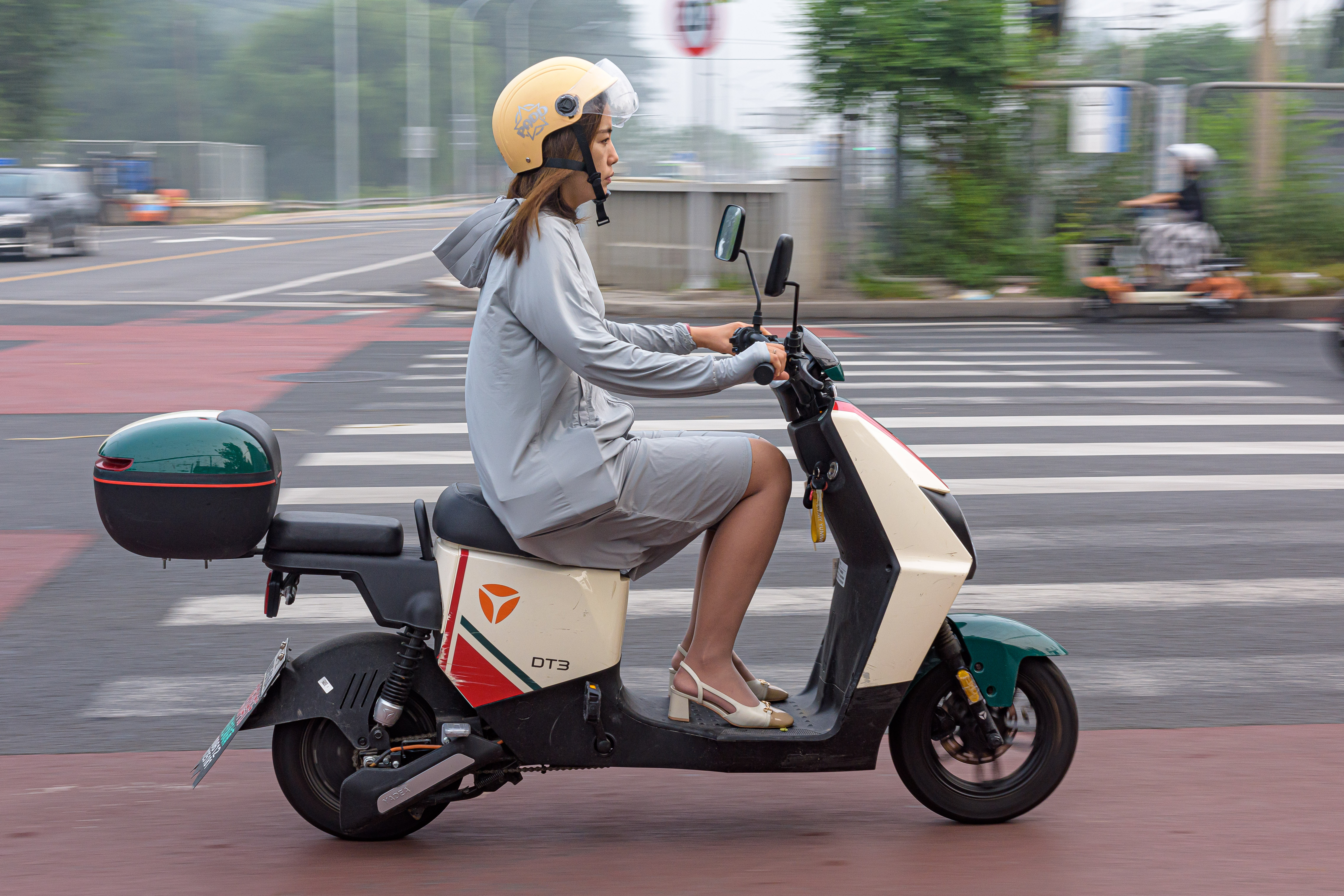 Woman in yellow helmet rides a green electric scooter at an intersection.