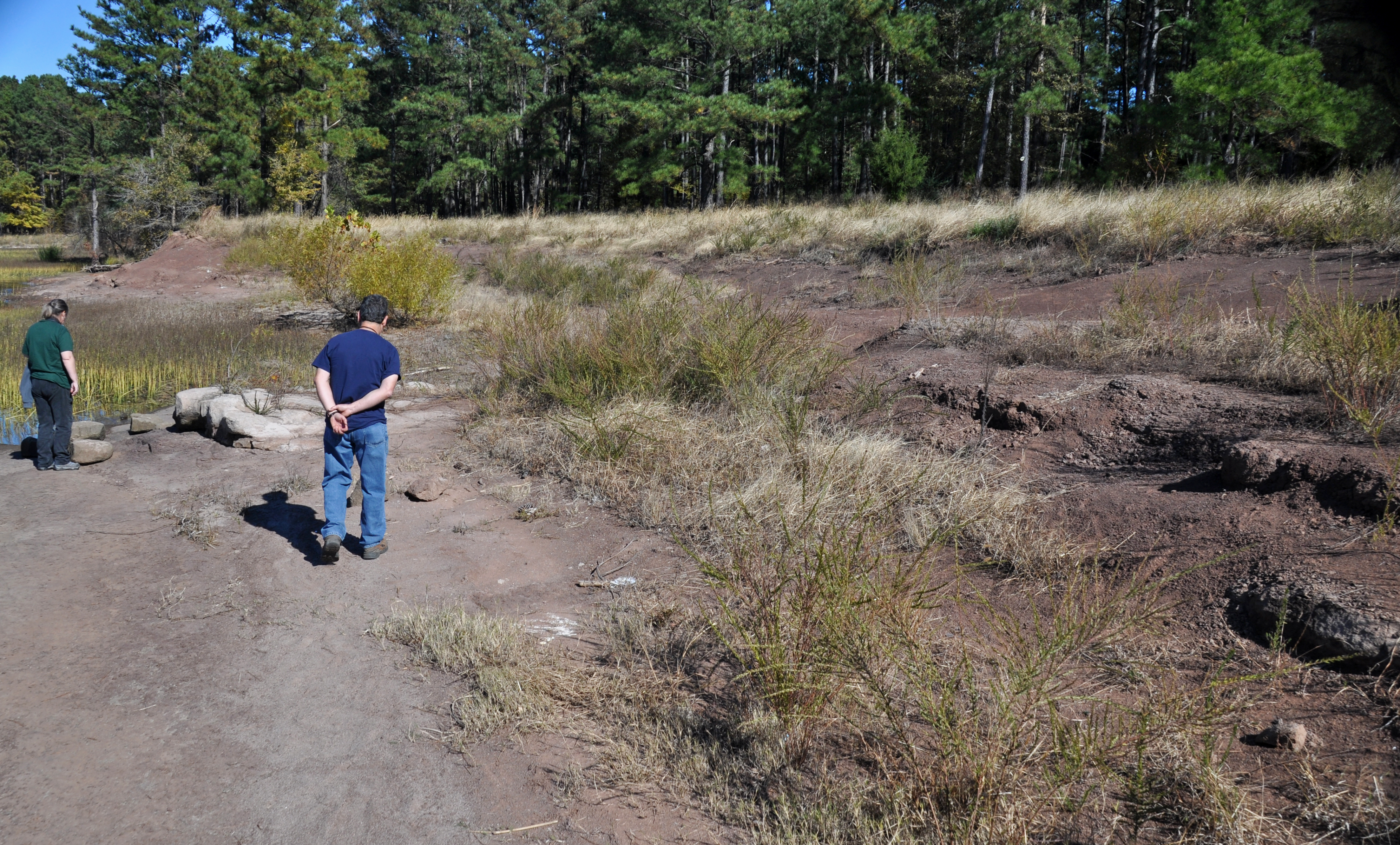 Two people stand on a wide, dry exposed lakebed with pine trees in the background.