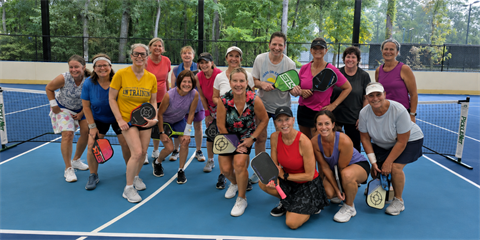 Group of smiling adults holding pickleball paddles on a blue outdoor court.