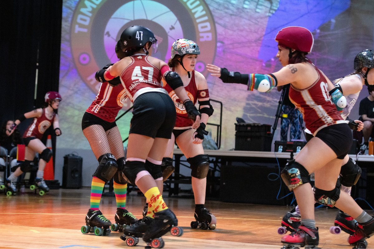 Roller derby players in red and black uniforms skate on a wooden floor during a match.