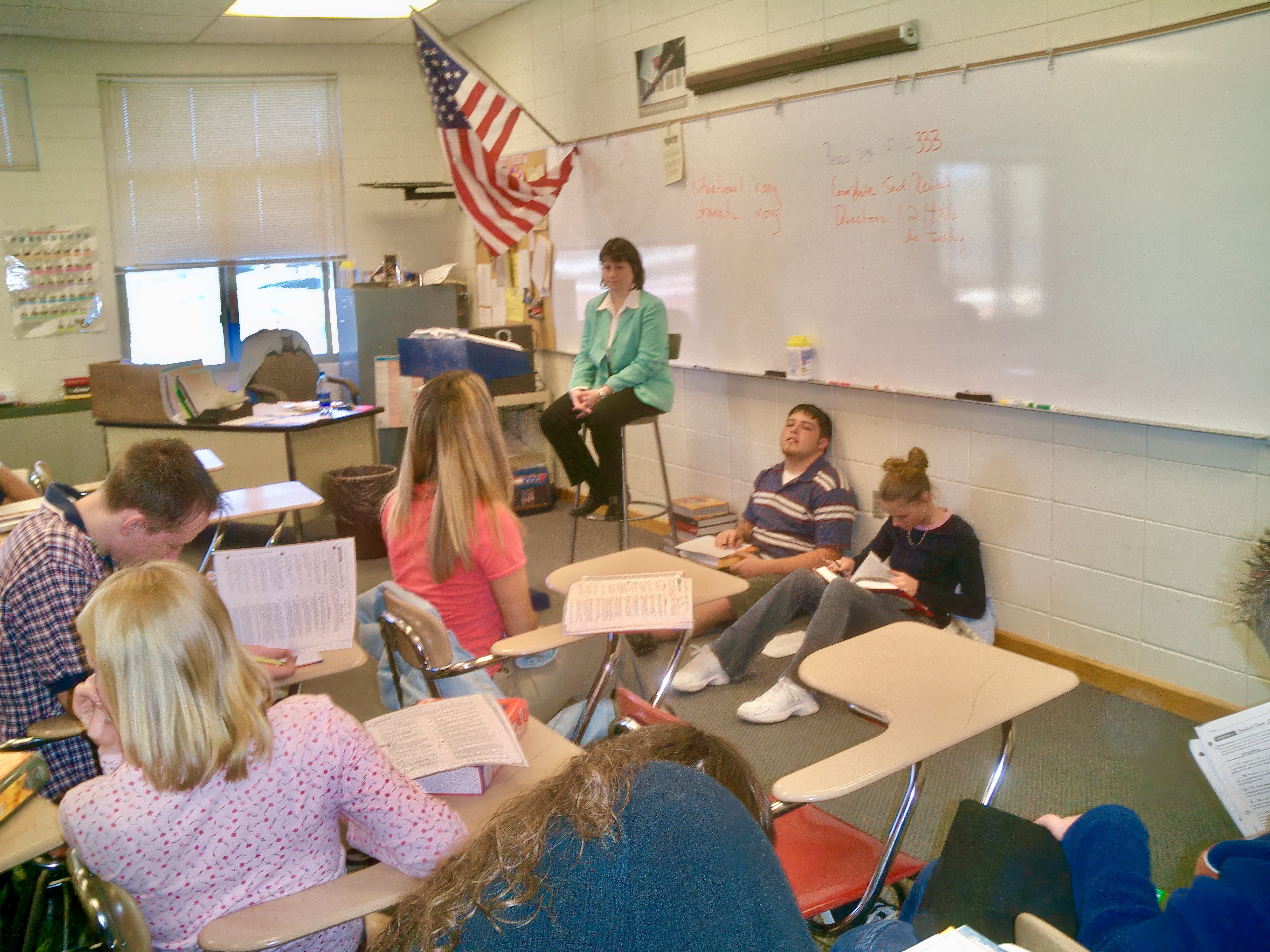 A teacher sits at the front of a classroom with students at desks.