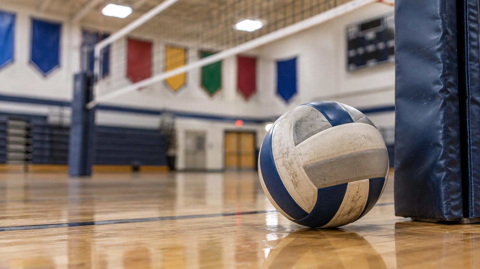 A volleyball resting on a hardwood gym floor next to a net post.