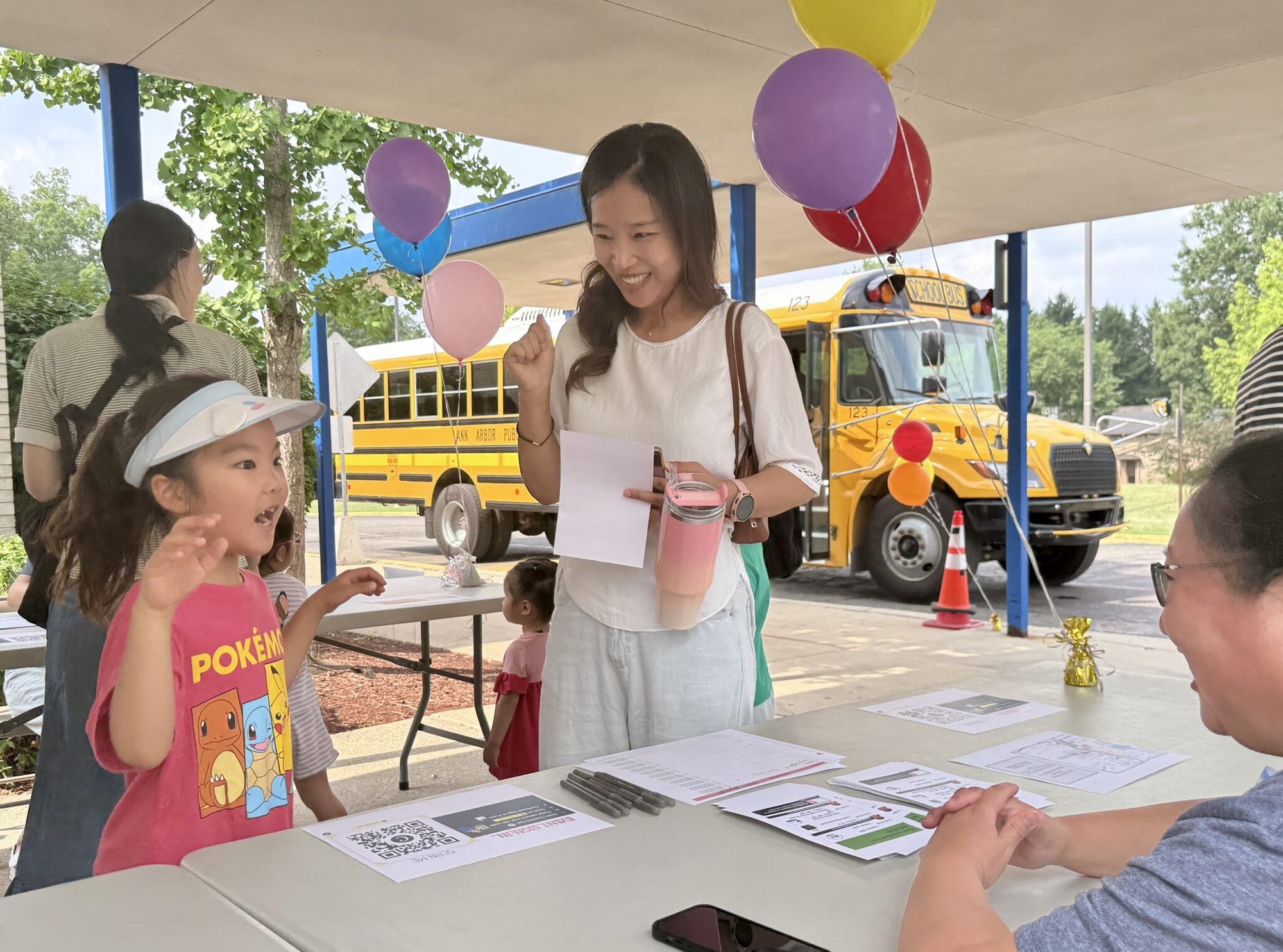 A woman and young girl stand at an outdoor table under a tent with balloons and a school bus behind them.