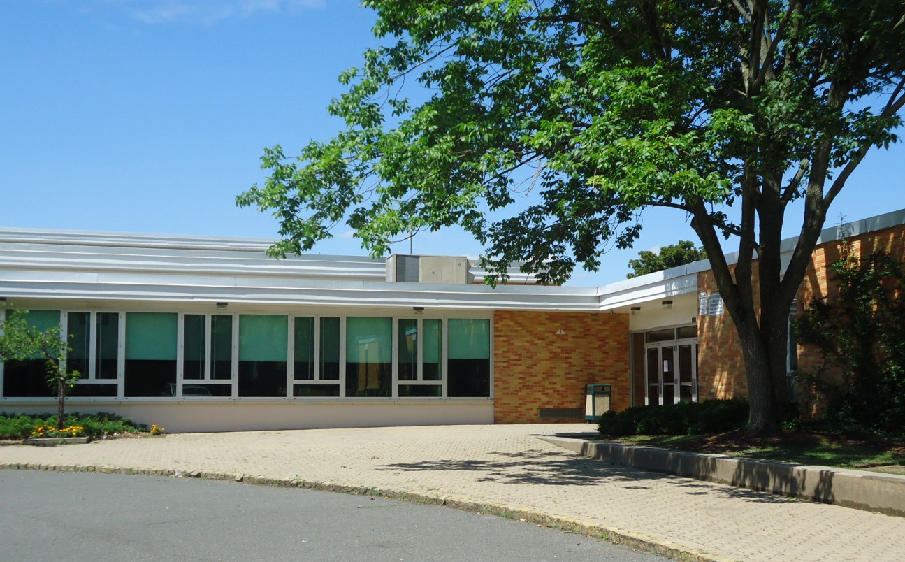 Brick school building entrance with a paved driveway and large tree out front.
