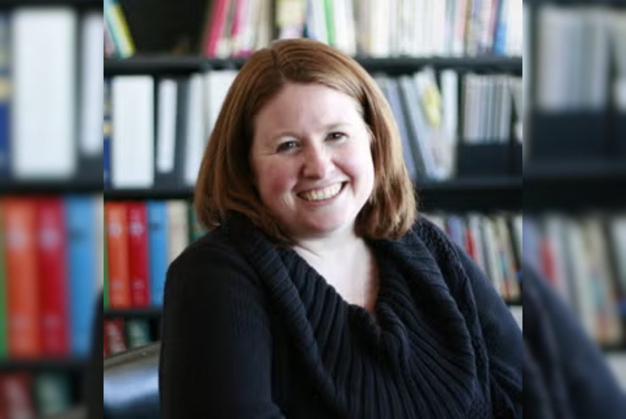 Smiling woman with reddish-brown hair seated in front of a bookshelf.