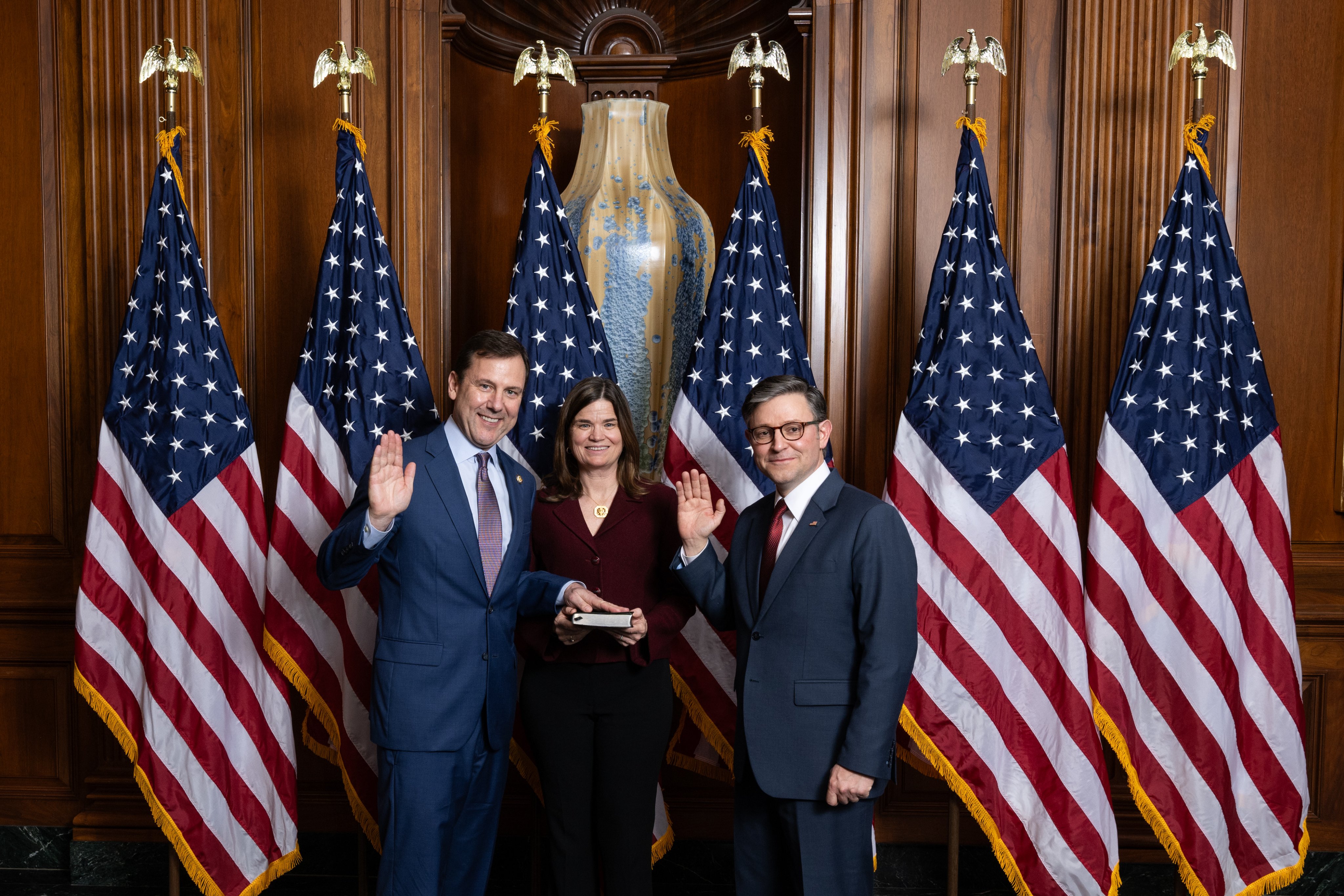 Three people stand before American flags in a wood-paneled congressional chamber, two with right hands raised.
