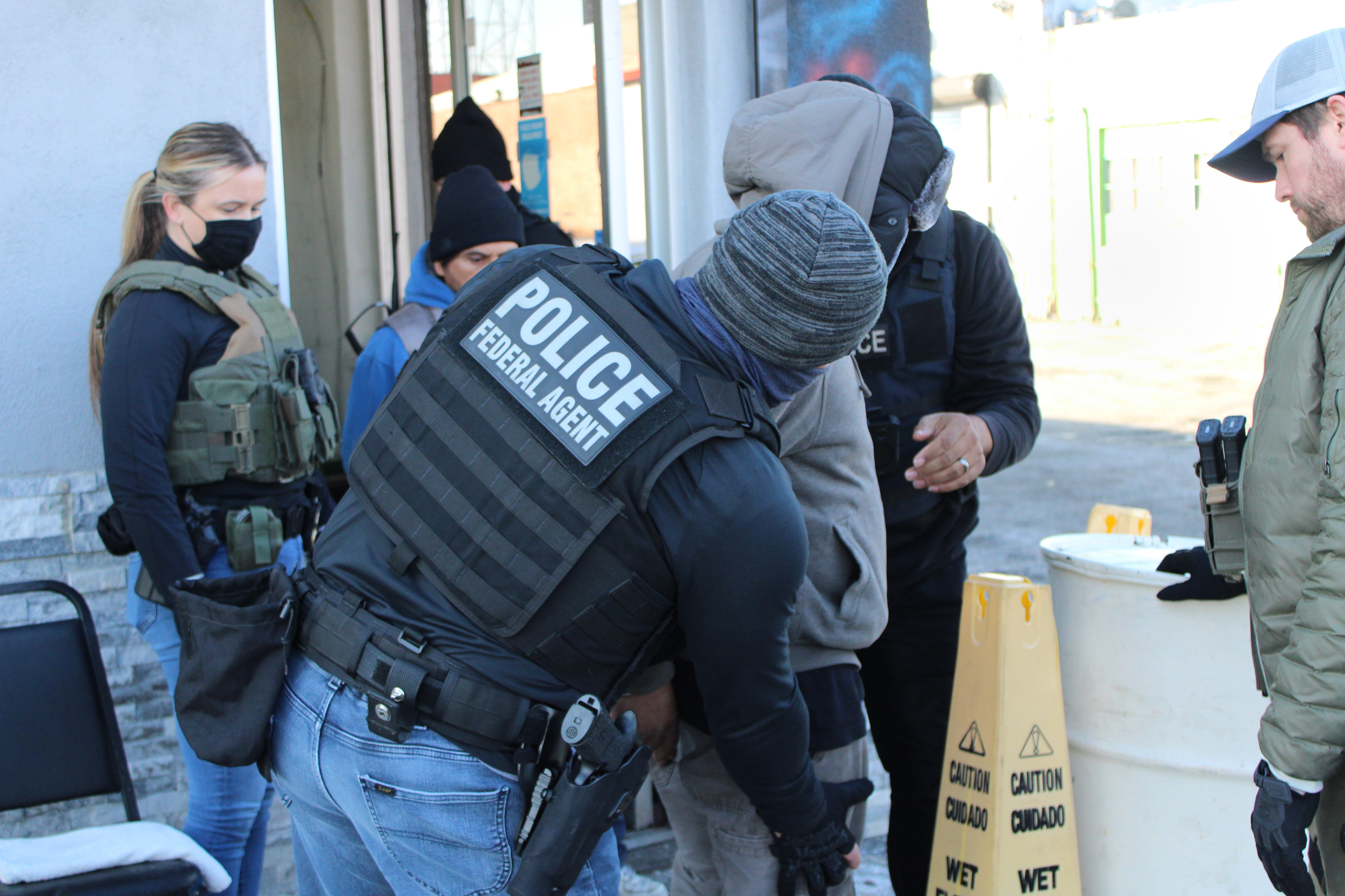 Federal agents in police vests detain a person outside a building during an enforcement operation.