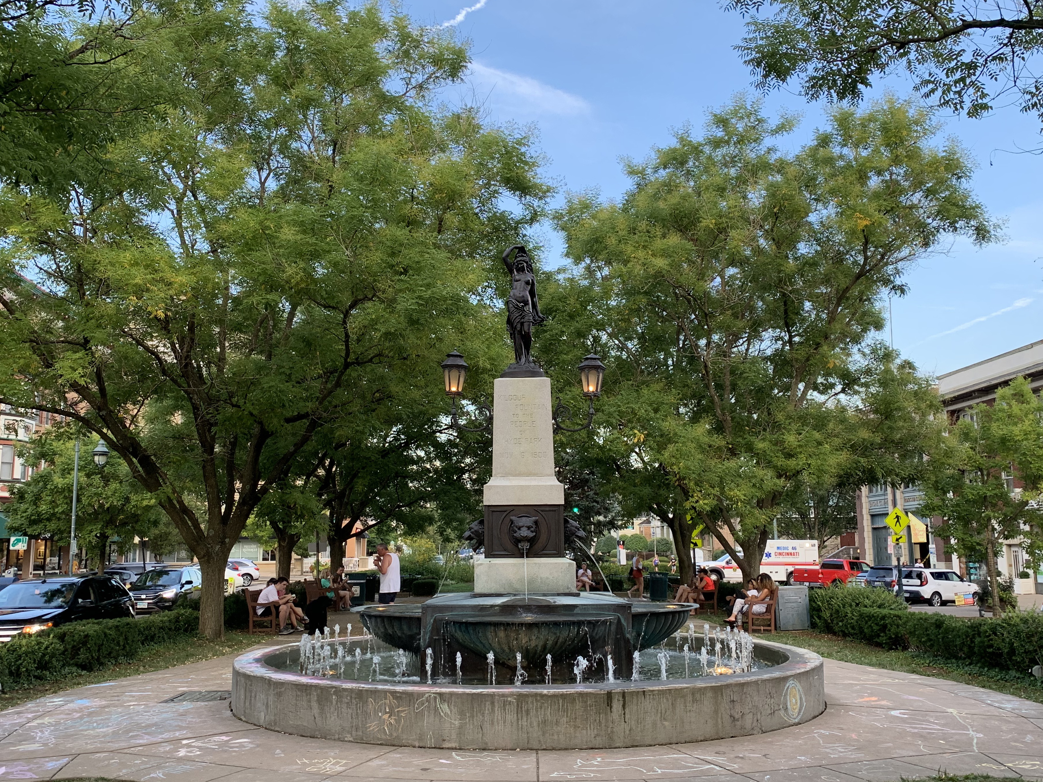 Circular fountain with bronze statue and lamp posts surrounded by green trees in a public square