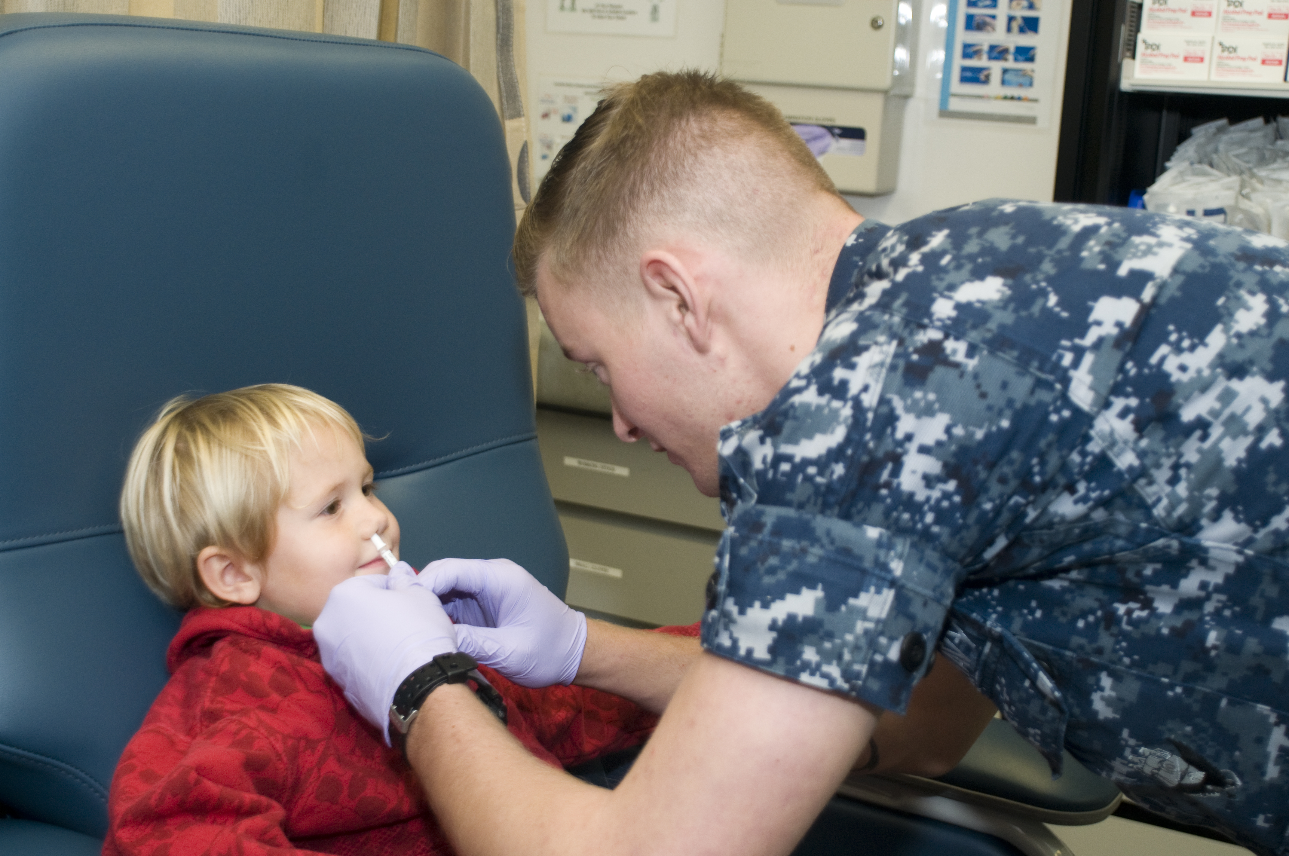 A healthcare worker administers a nasal spray flu vaccine to a young child in a clinic chair.