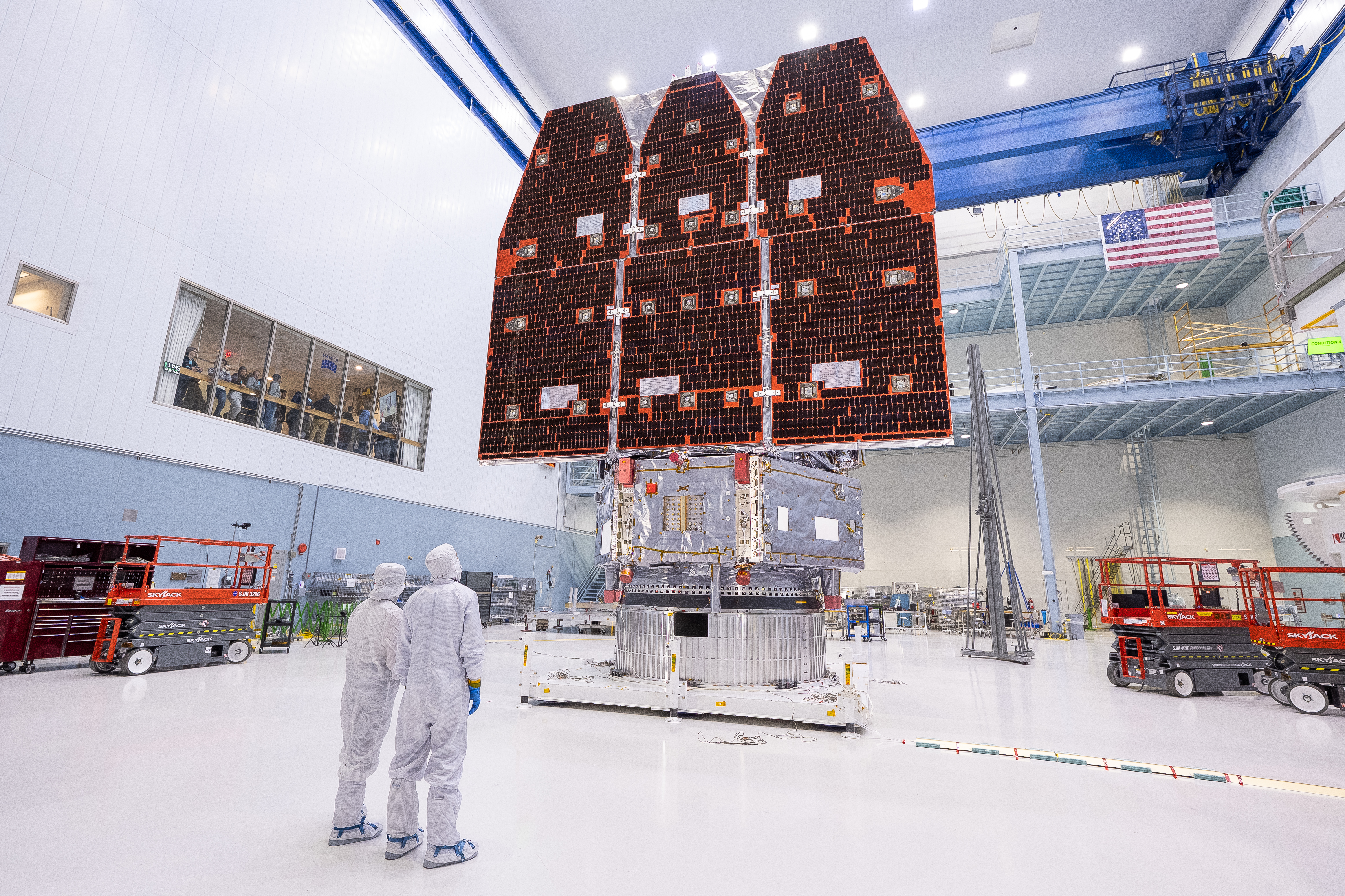 Two technicians in white cleanroom suits look up at a large spacecraft with deployed solar panels.