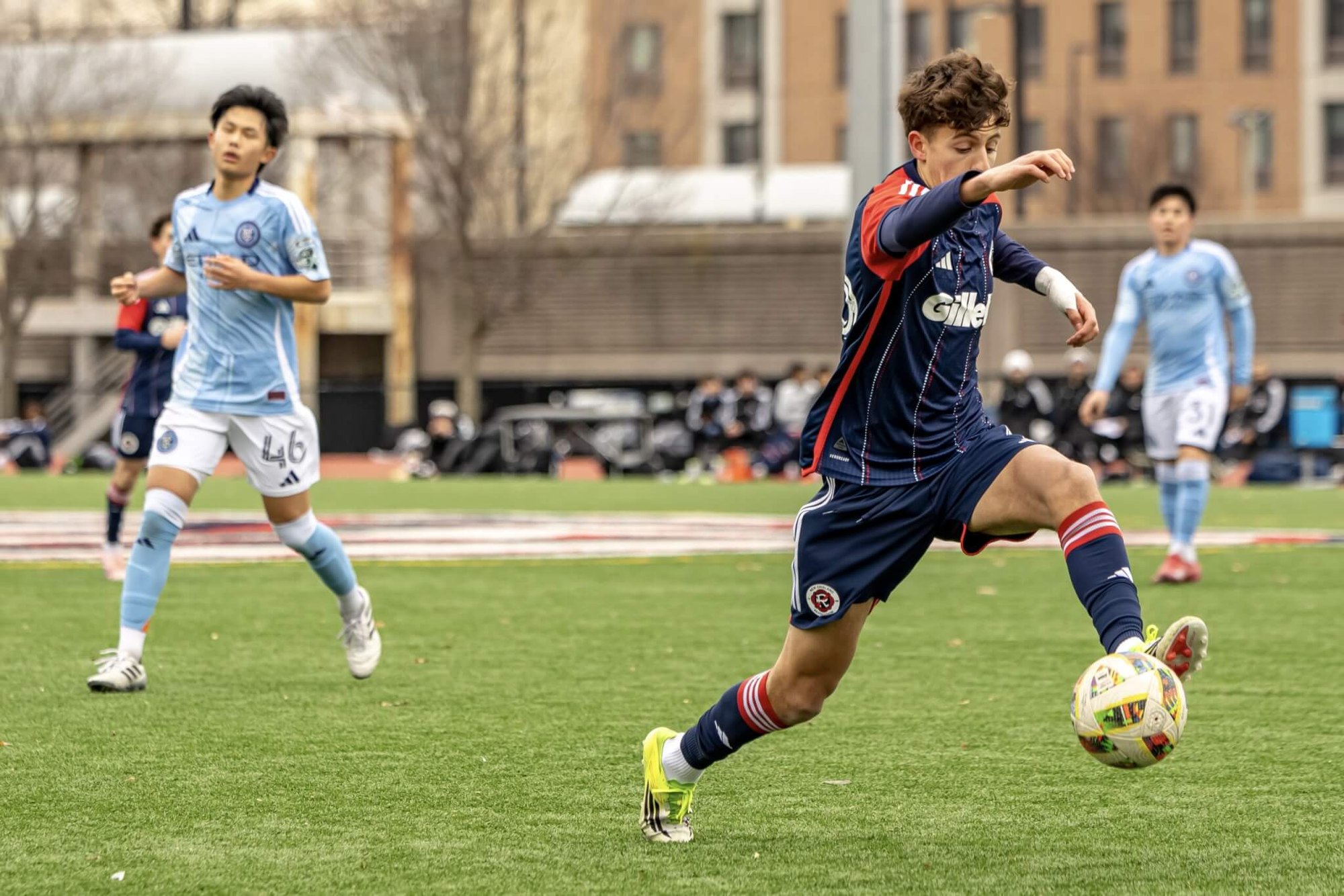 A youth soccer player in a navy Revolution kit dribbles past opponents in light blue uniforms on a grass field.